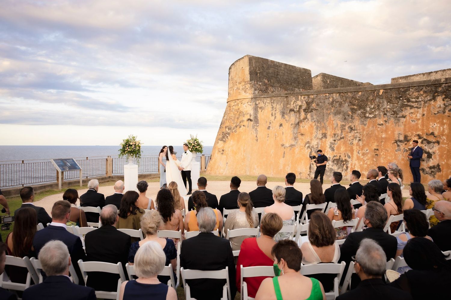 Wedding ceremony overlooking historic fortress walls with guests seated facing ocean view.