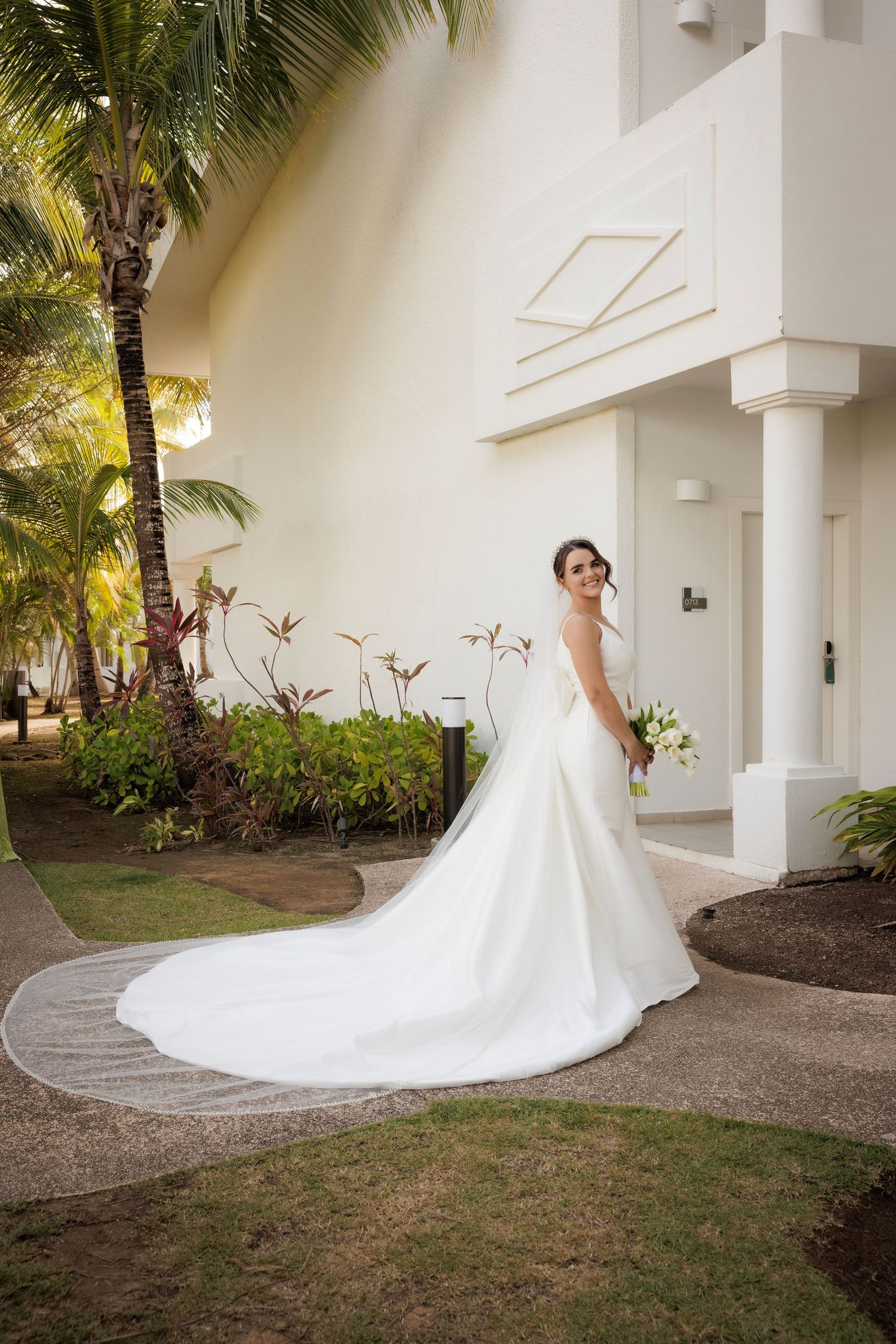 A stunning white wedding gown with a long train cascades across tropical garden path with palm trees and white columns.