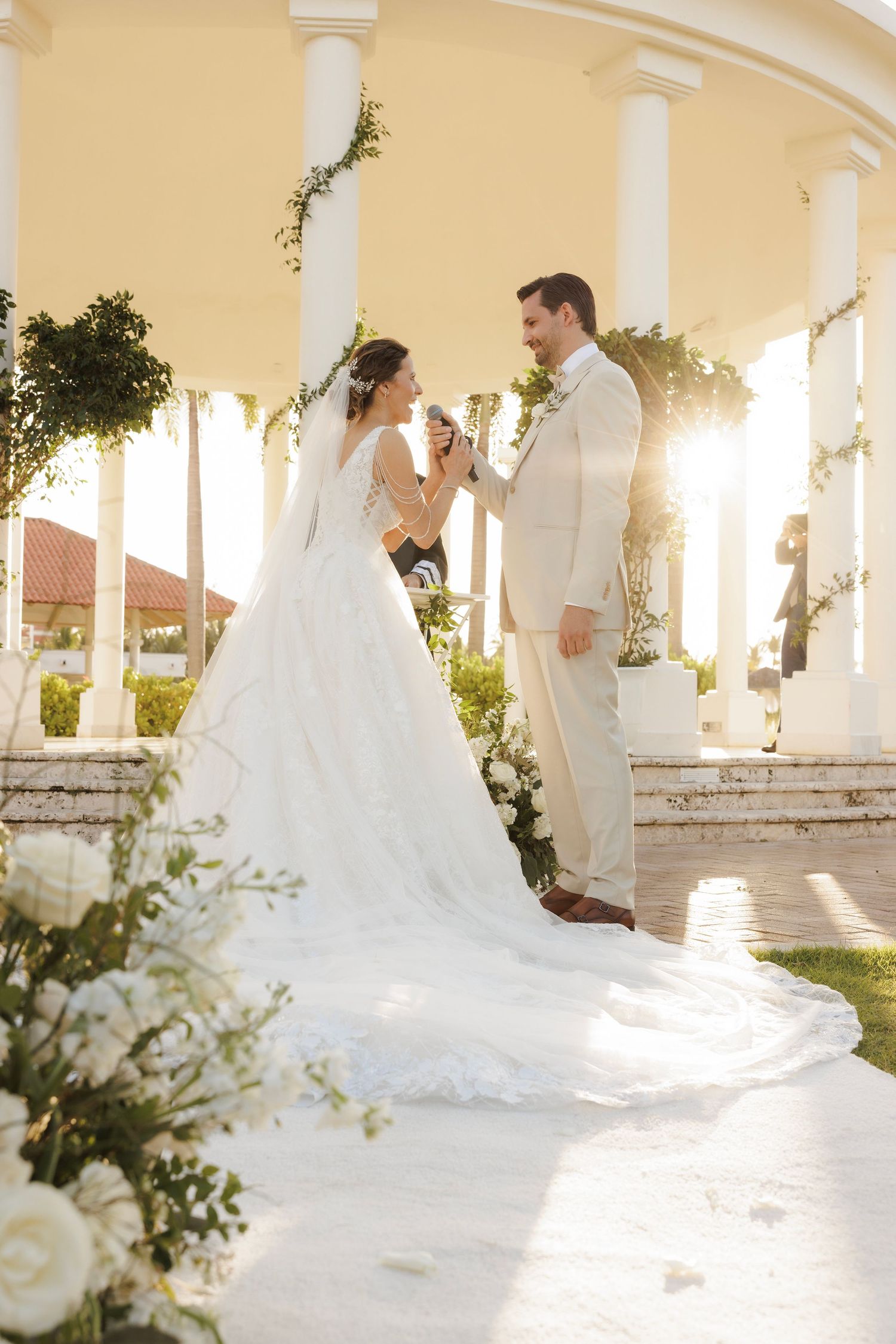 A beautiful wedding couple stands in a sunlit gazebo with white columns and flowing bridal gown with long train.