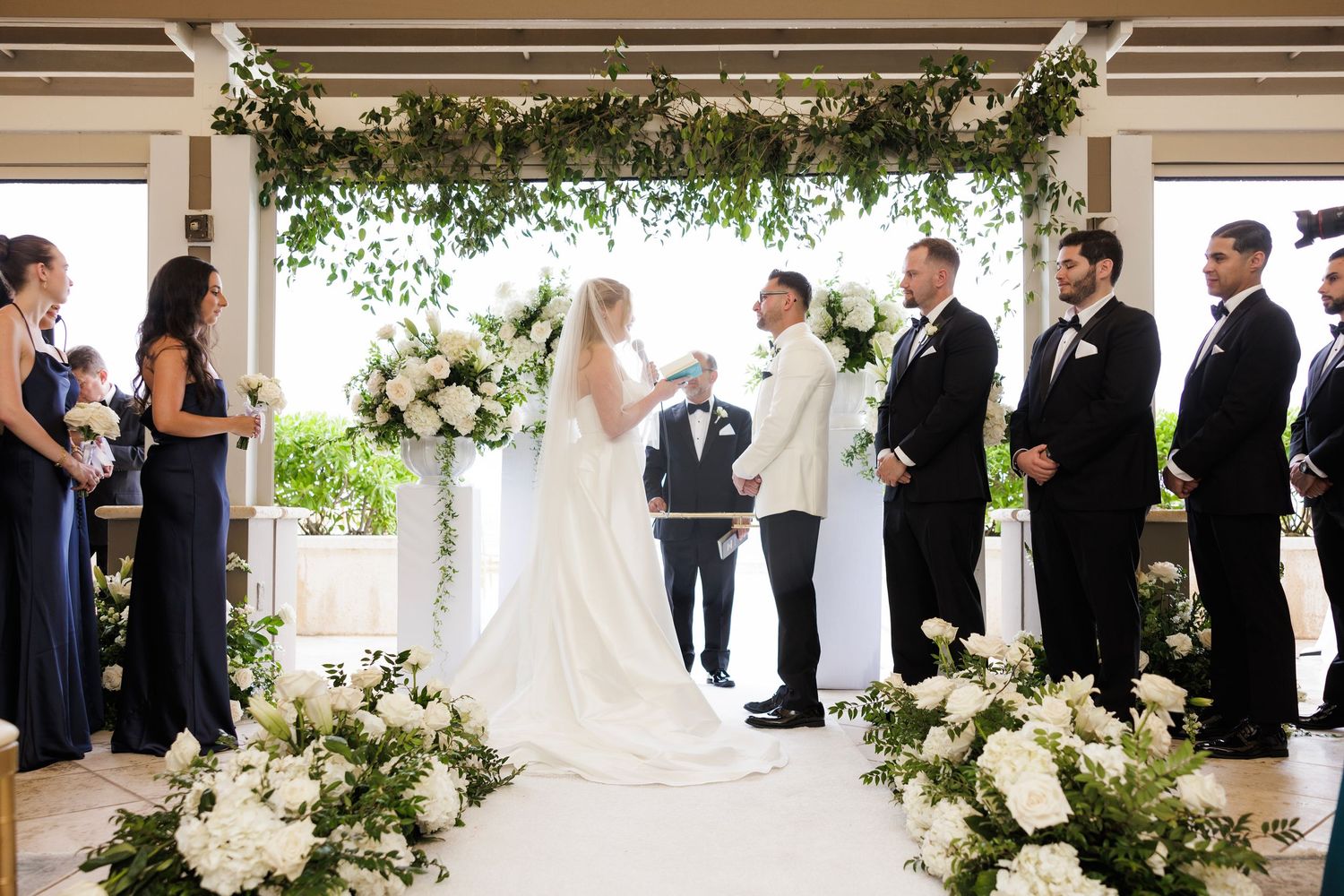 Elegant wedding ceremony setup with white floral arrangements and greenery in an outdoor covered space.