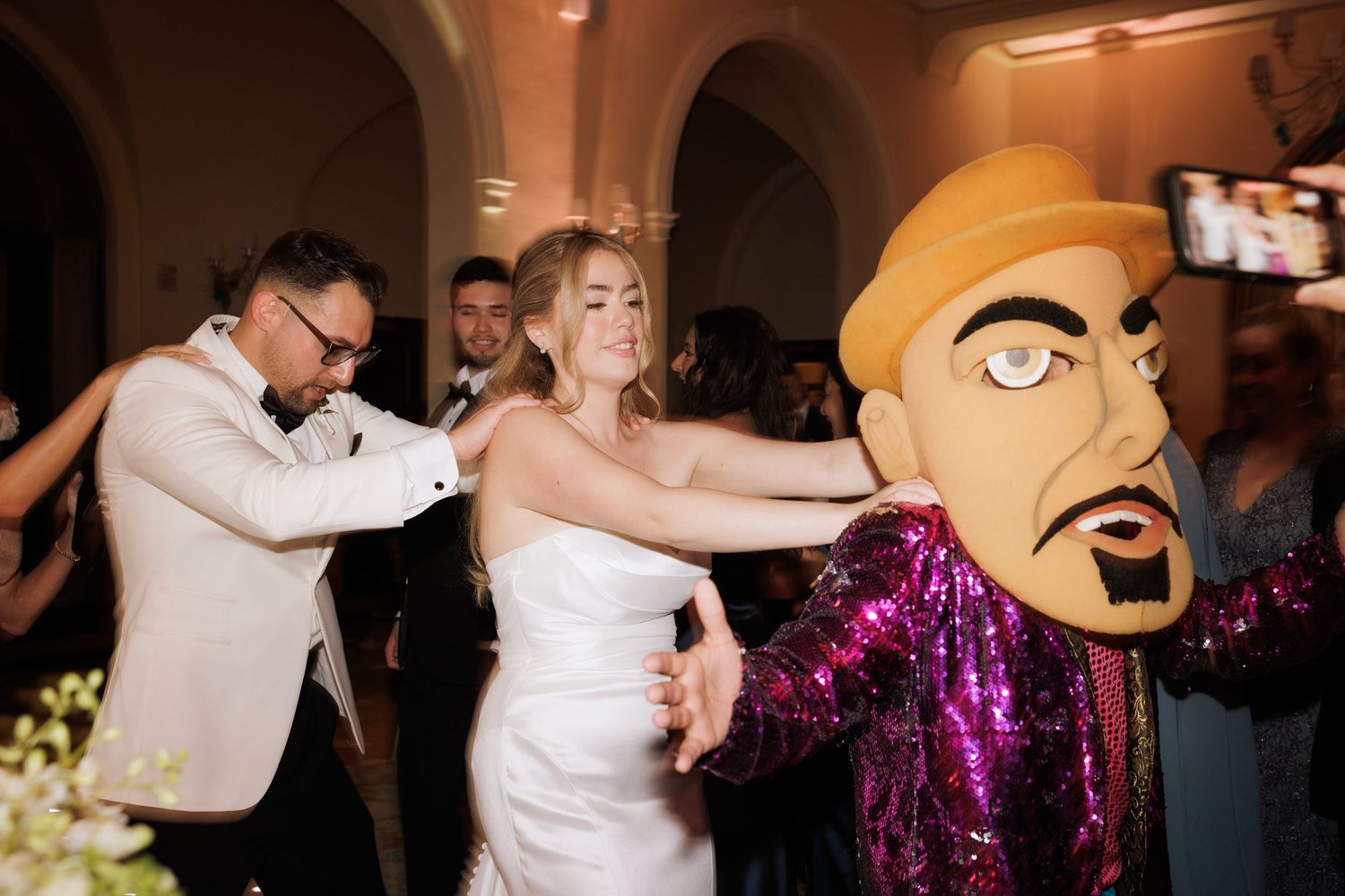 Wedding guests dance with a person wearing a large puppet head mask and sparkly purple jacket at a reception celebration.