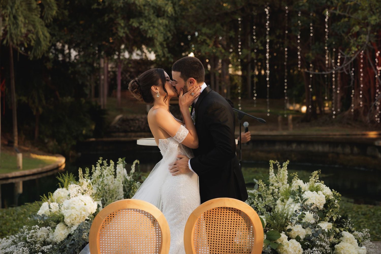 Romantic outdoor wedding scene with cane back chairs and elegant white floral arrangements beside a pond at sunset.