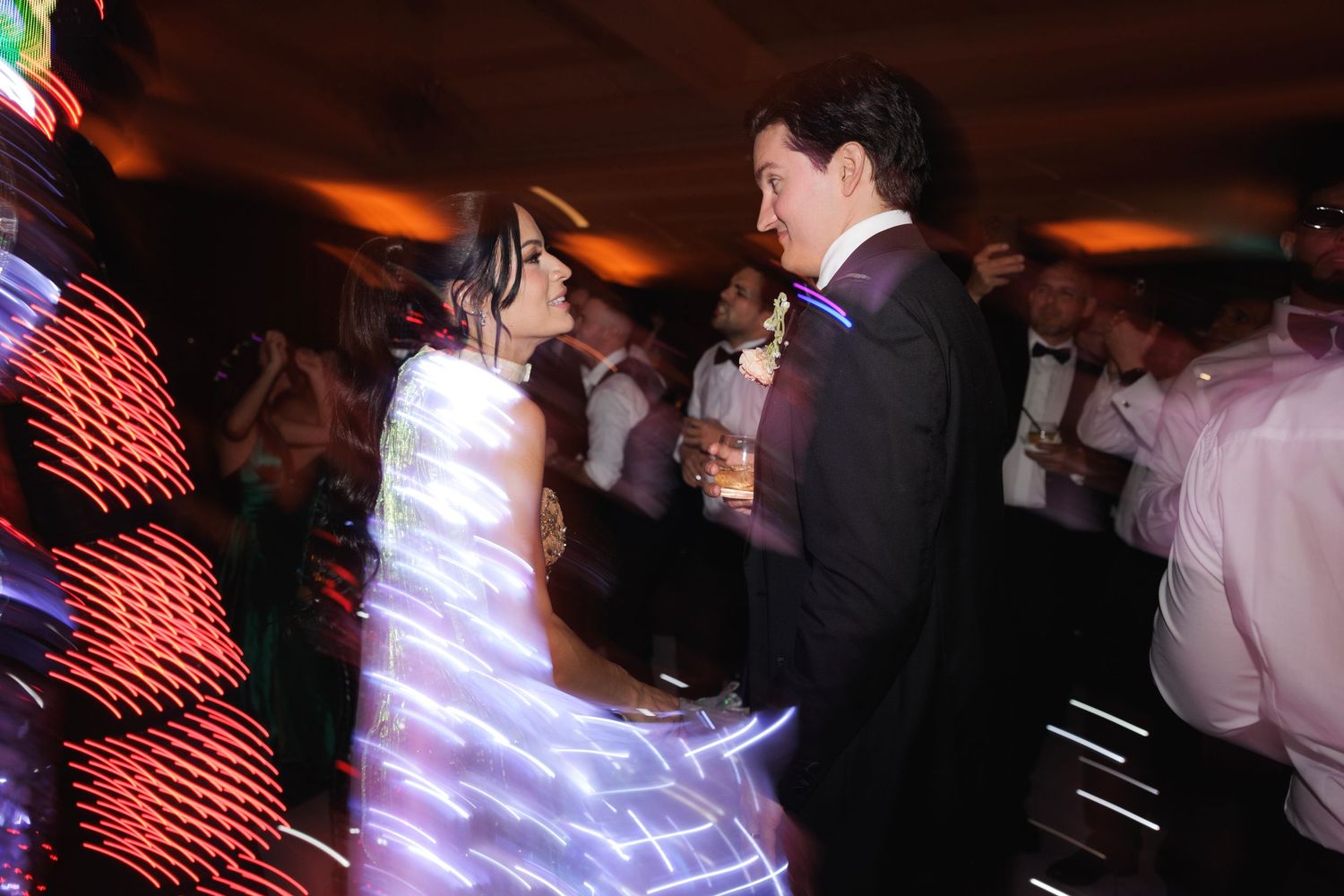People dance and socialize at a dimly lit nightclub or party venue with glowing lights in the background.