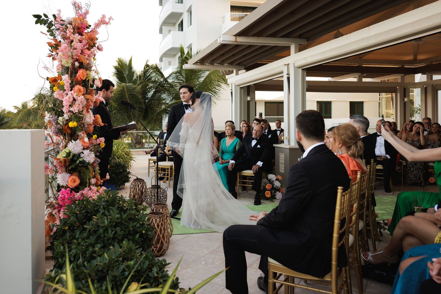 A greenhouse wedding ceremony with pink floral arrangements and guests seated in white chairs.