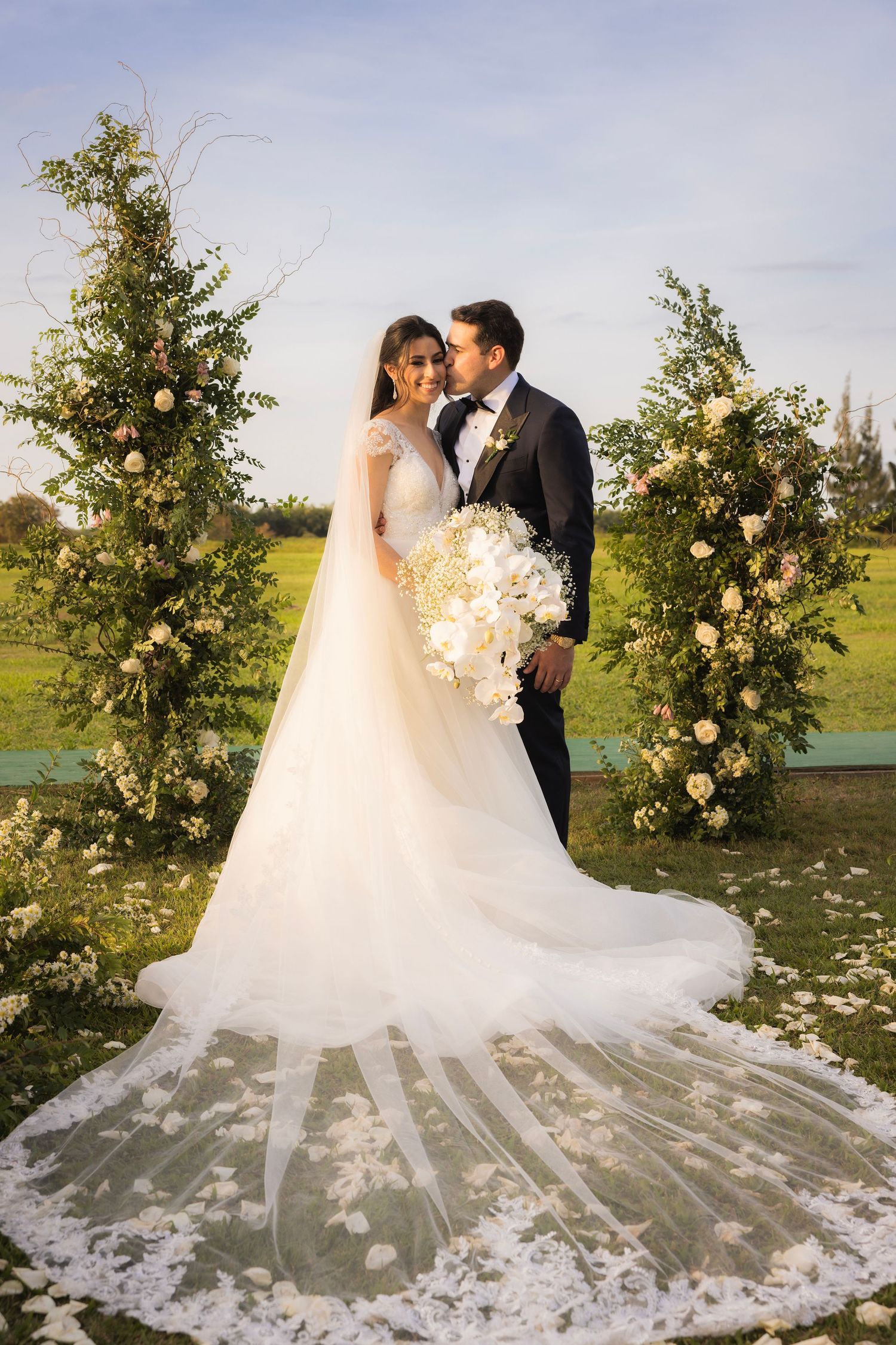 Elegant wedding couple poses by floral arch with bride's dress train spreading beautifully across green lawn.