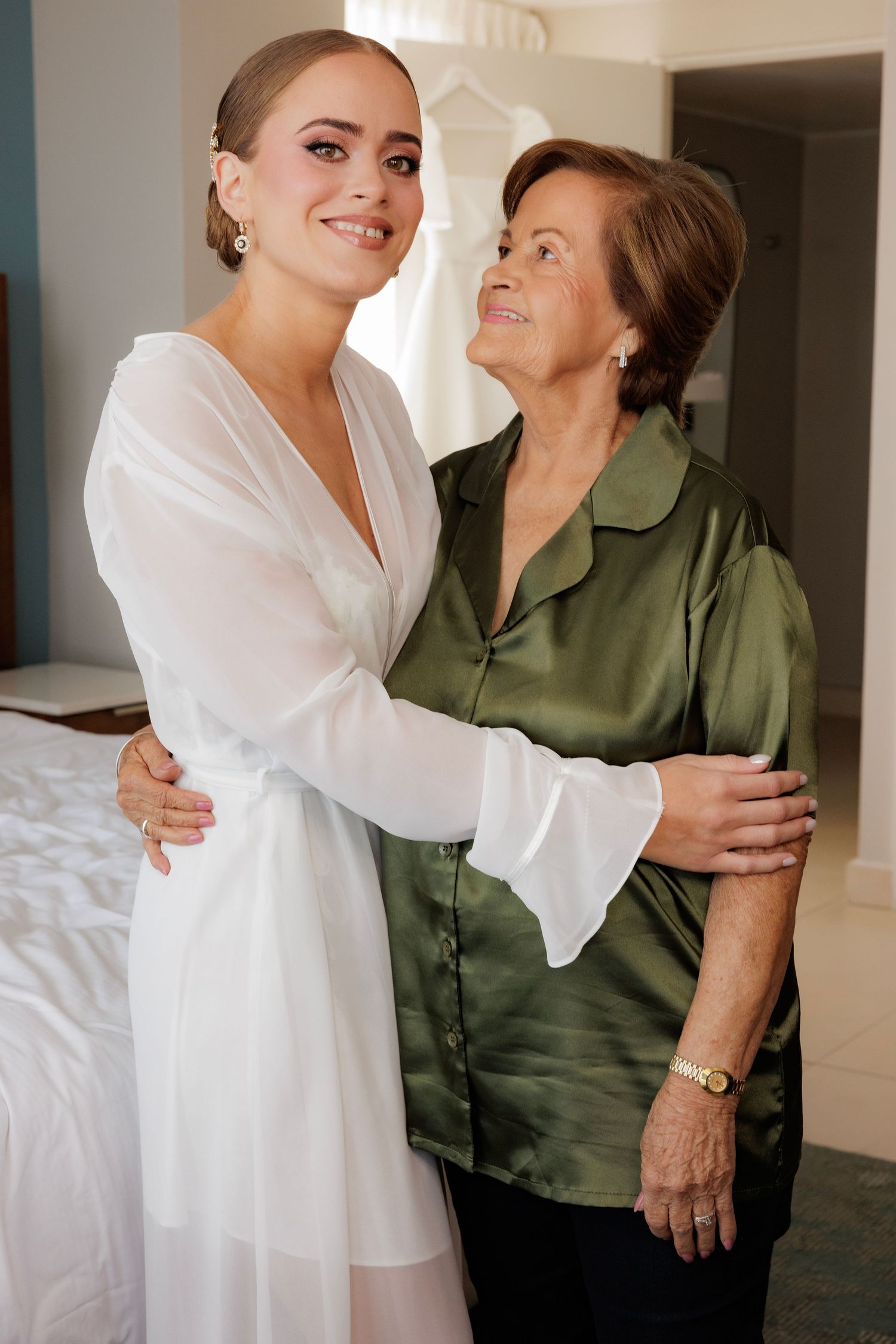 Two people embrace warmly, one wearing a crisp white outfit and the other in an olive green blouse.