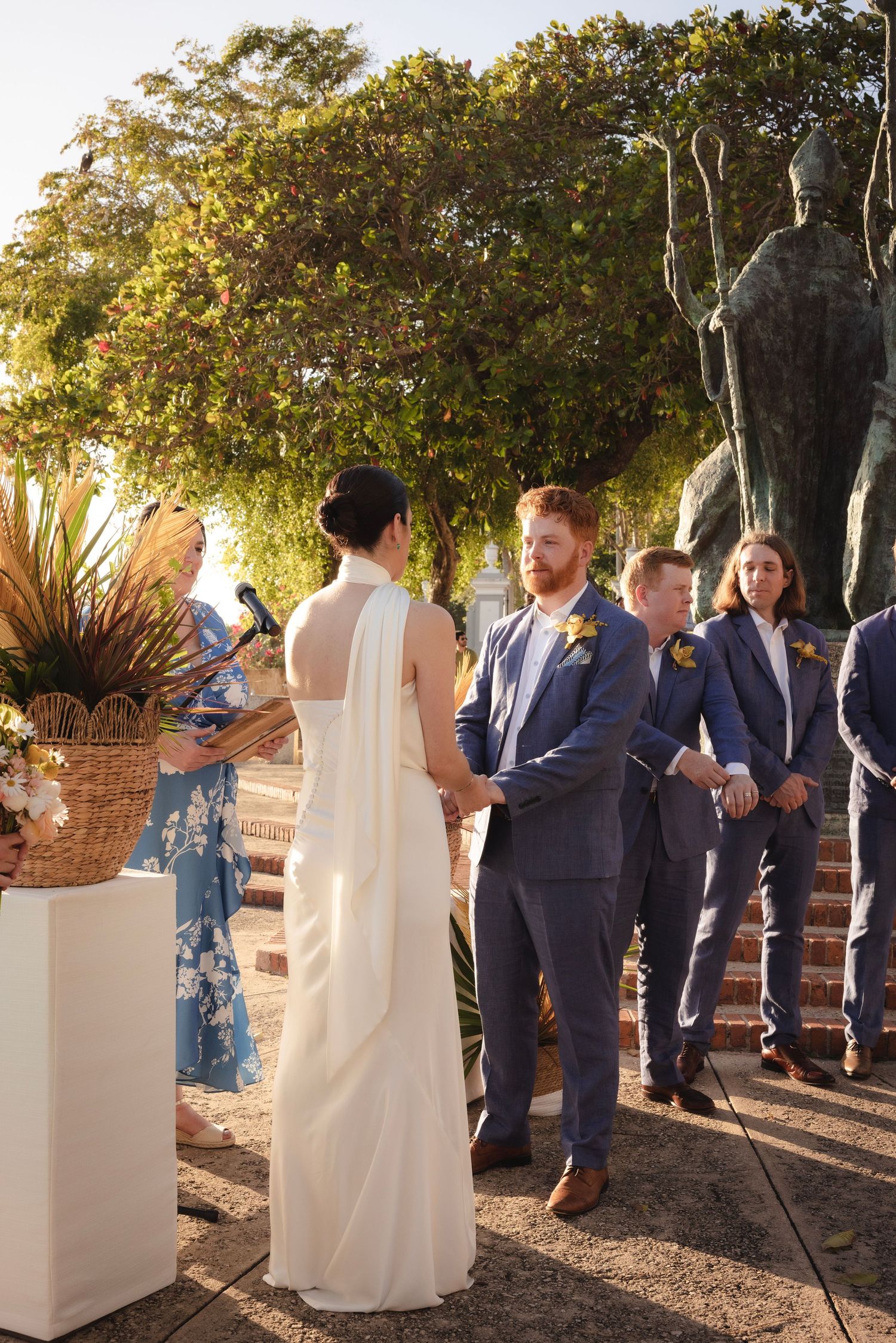A wedding ceremony taking place outdoors with guests in navy suits and a flowing white dress against a natural backdrop.