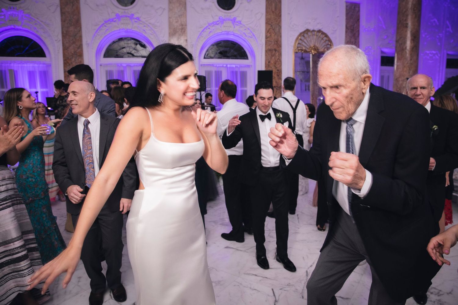 A group of guests dance and celebrate at an elegant wedding reception with purple uplighting in a grand ballroom.