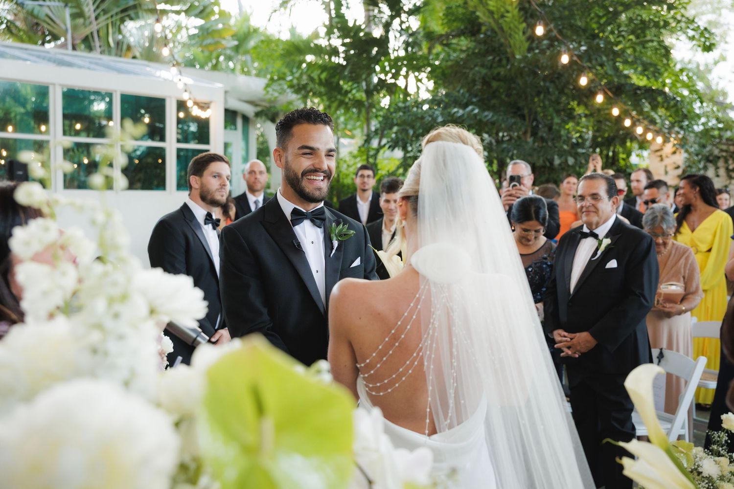 A joyful outdoor wedding ceremony takes place under string lights with white floral decorations and guests watching.