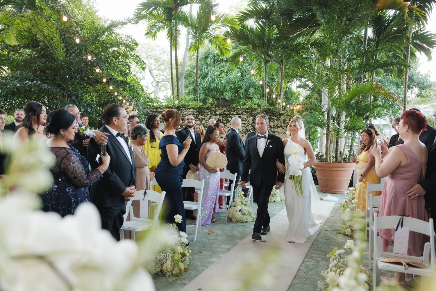 An outdoor wedding ceremony takes place in a lush tropical garden setting with palm trees and potted plants.