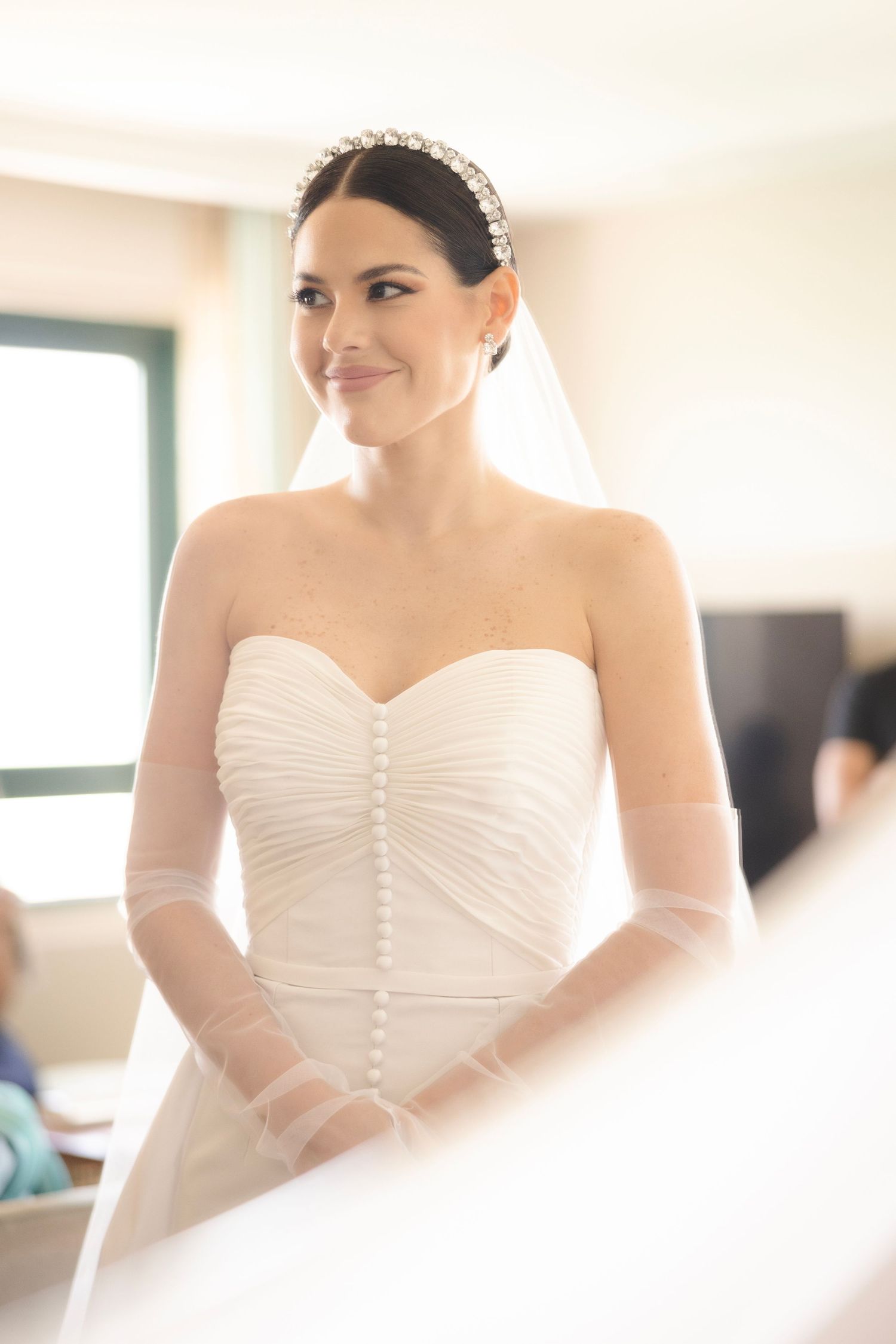 A bride wearing a classic white strapless wedding gown with ruched bodice and sheer long sleeves stands elegantly with a pearl headpiece.