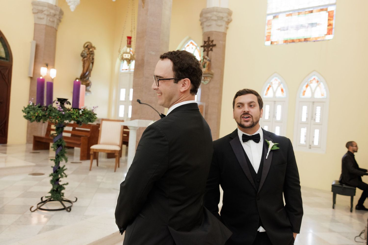 Two men in formal black tuxedos stand in an elegant church interior with arched windows and candles.