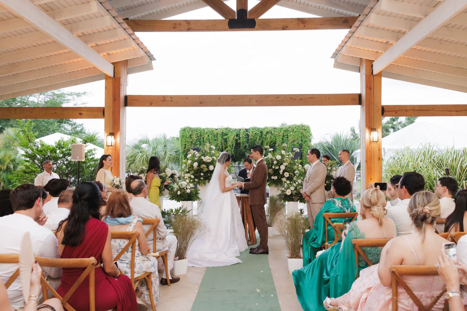 An outdoor wedding ceremony under a covered pavilion with guests seated on white chairs and green decorations.