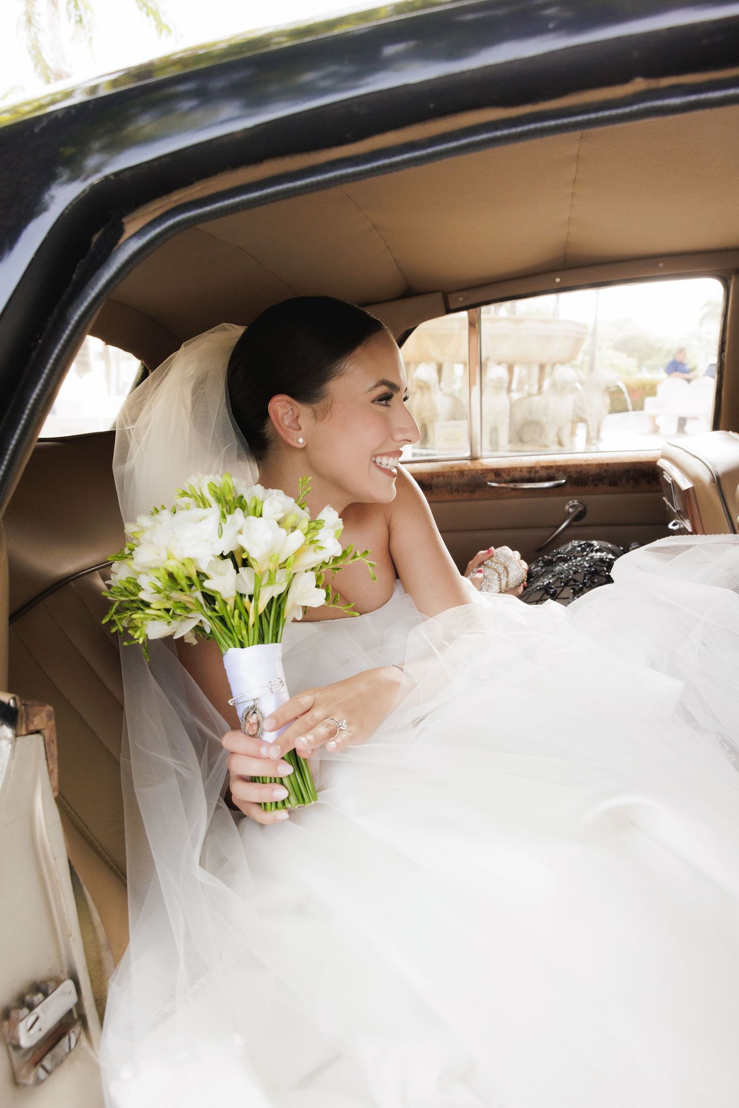 Bride in white dress holding bouquet of white flowers sits joyfully in vintage car on wedding day.