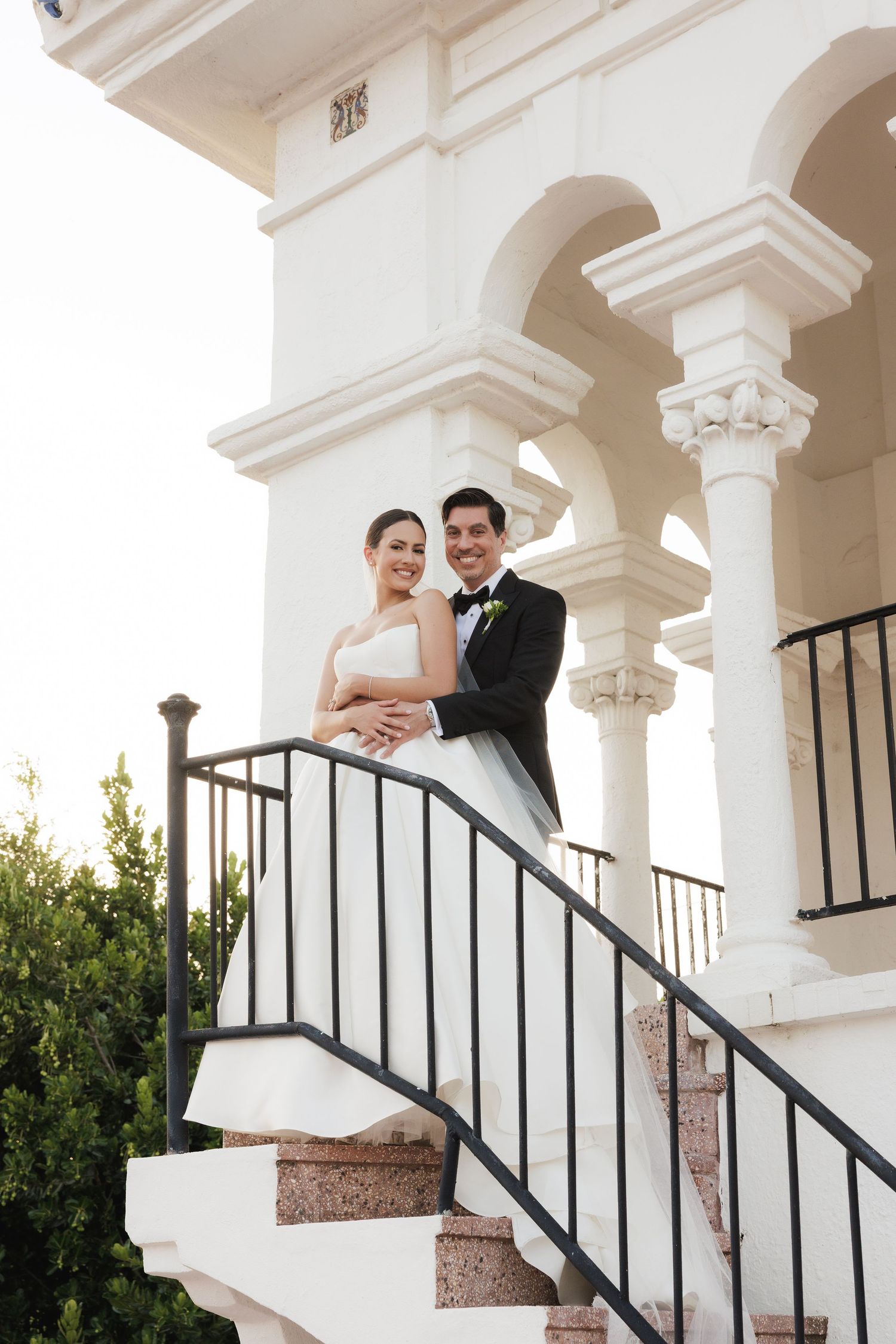A bride and groom pose on white stucco stairs with elegant archways and columns in the background.