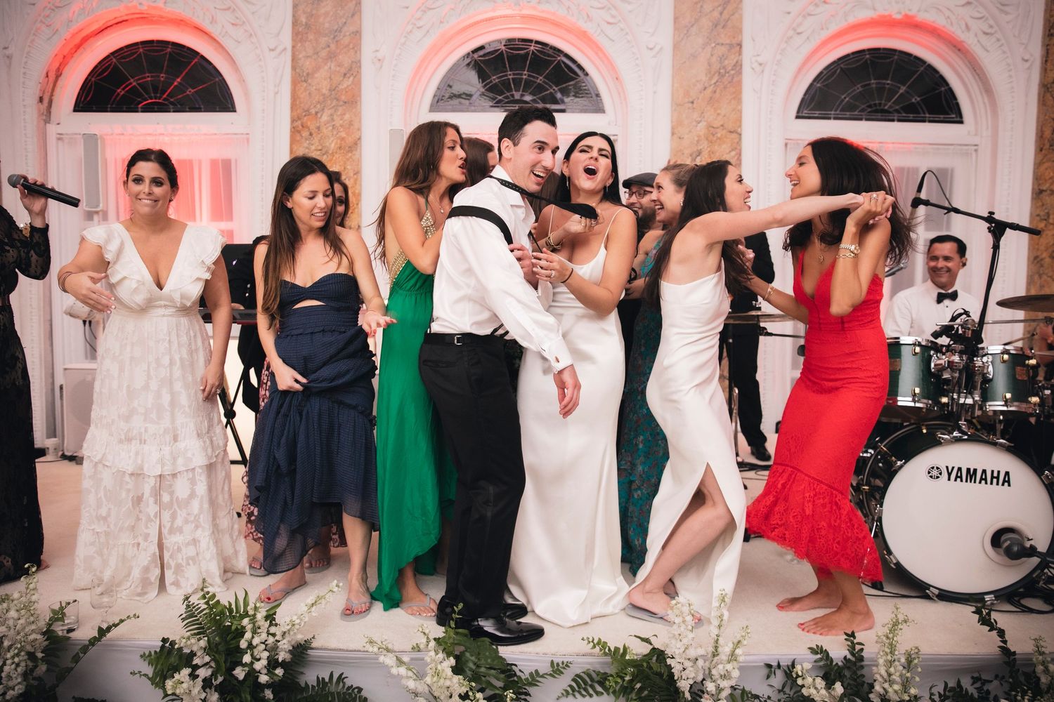 Wedding party and guests dancing together in a banquet hall with pink-tinted lighting and classical architecture.