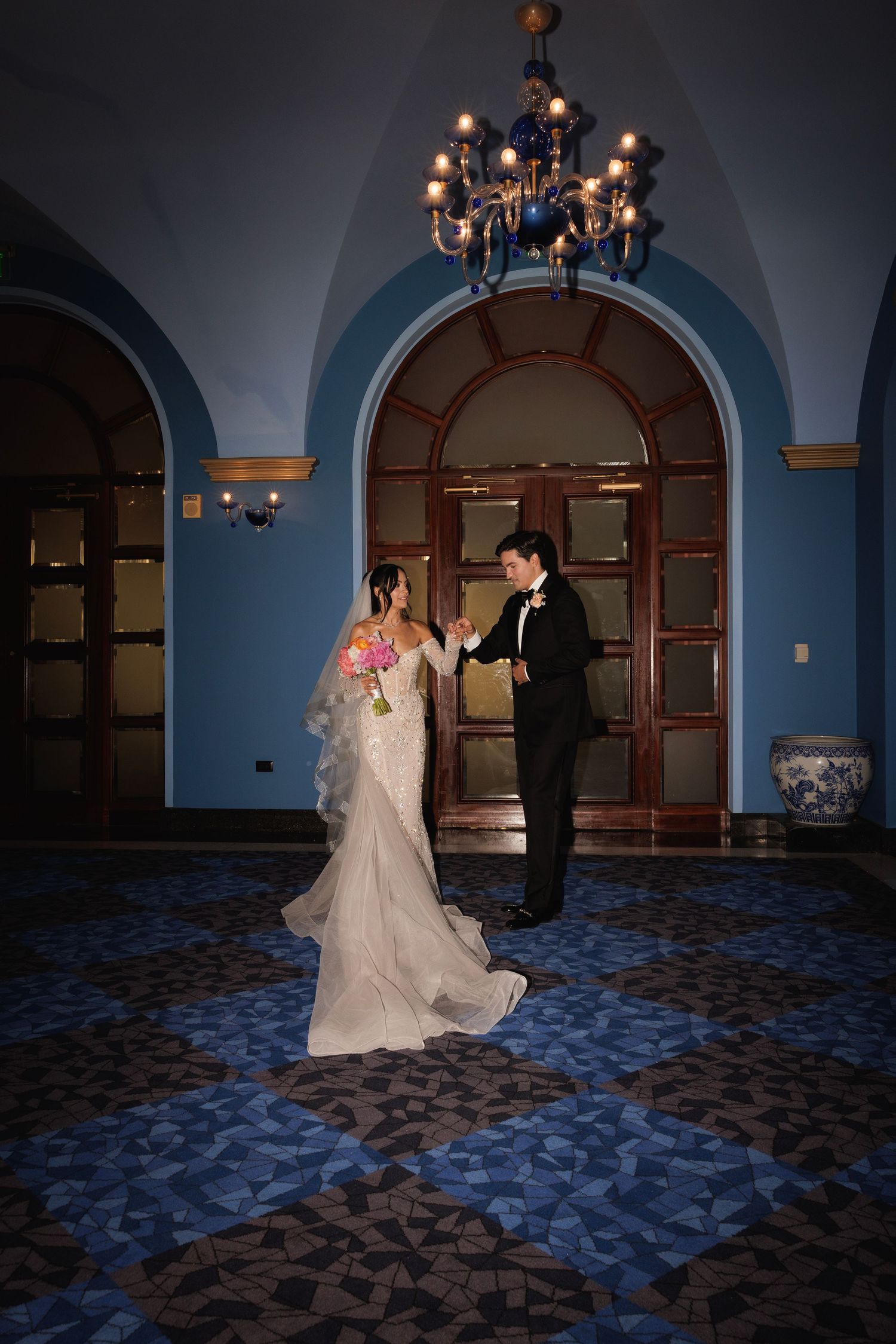 Couple dancing in an elegant ballroom with blue walls, arched doorways, and a decorative chandelier overhead.