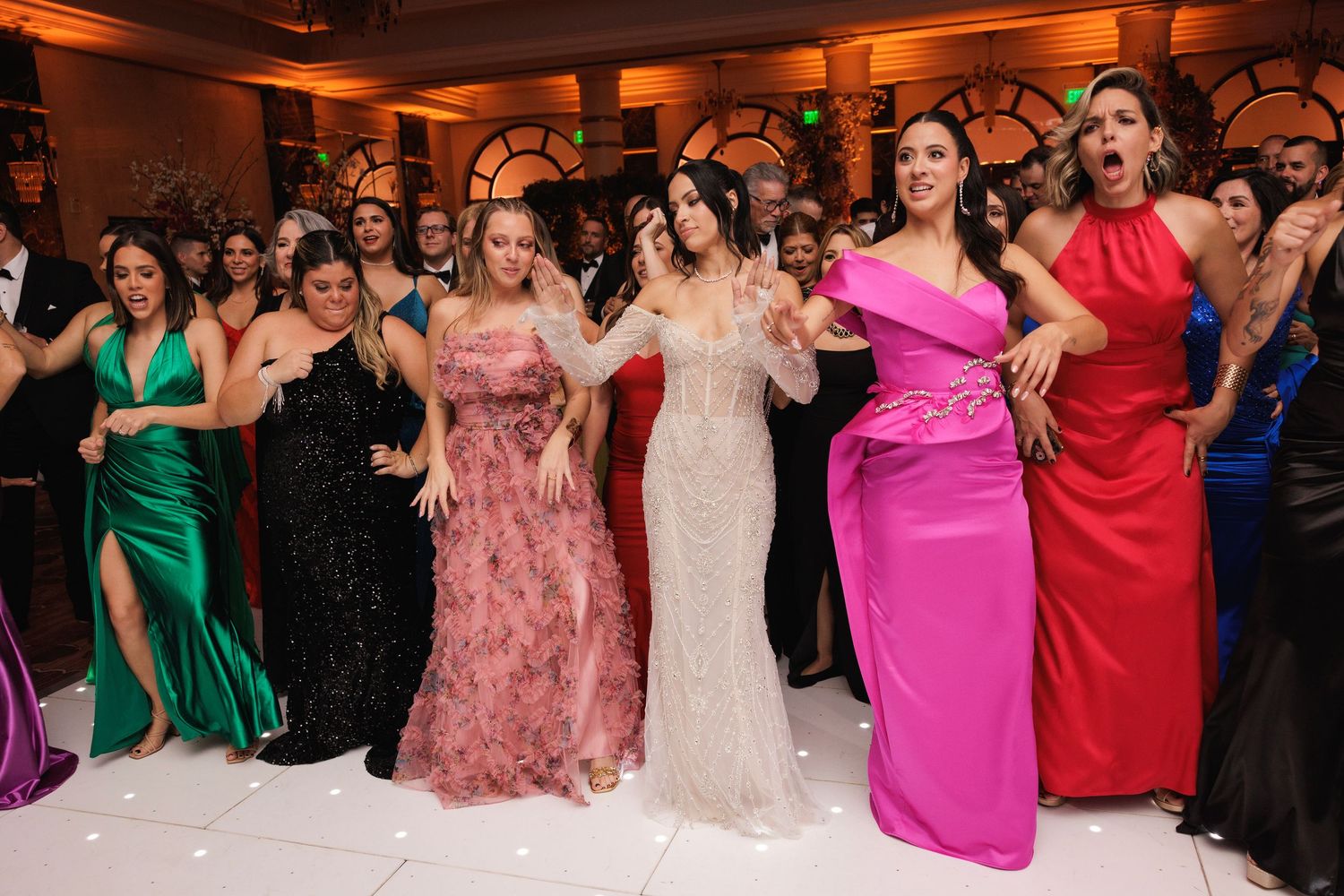 A group of wedding guests in elegant formal dresses in vibrant colors smile and pose together on a dance floor.