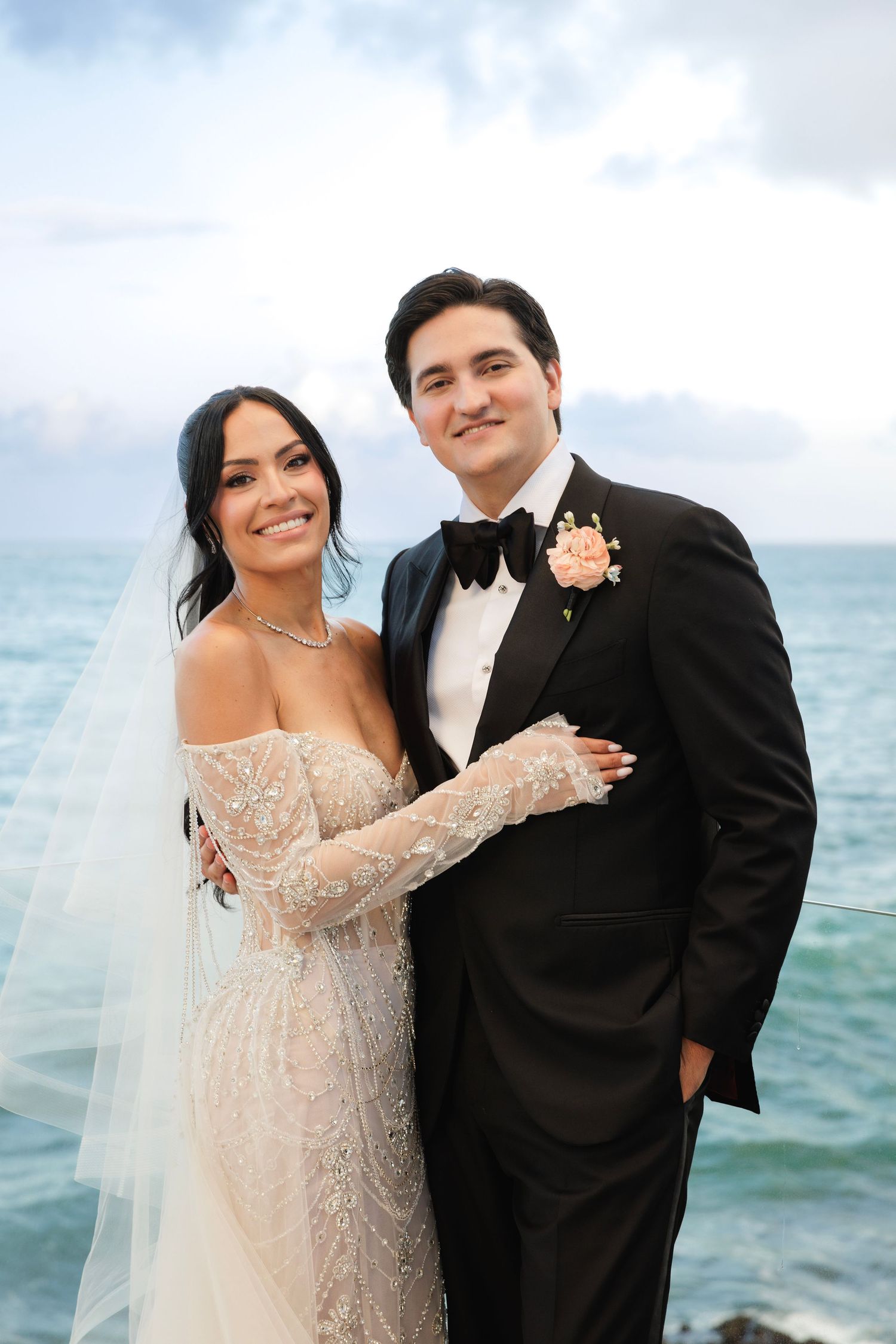 Elegant wedding couple poses against ocean backdrop with cloudy skies.