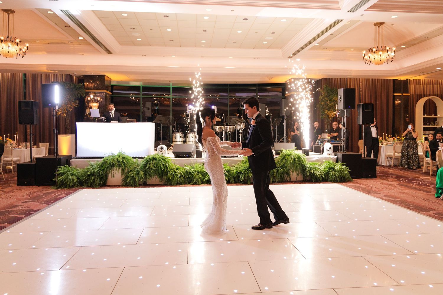 A couple shares their first dance on a white dance floor surrounded by indoor sparklers and elegant decor at a wedding reception.
