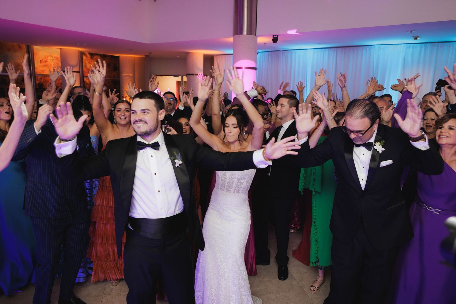 Large group of wedding guests filling dance floor while celebrating under dramatic purple lighting effects.