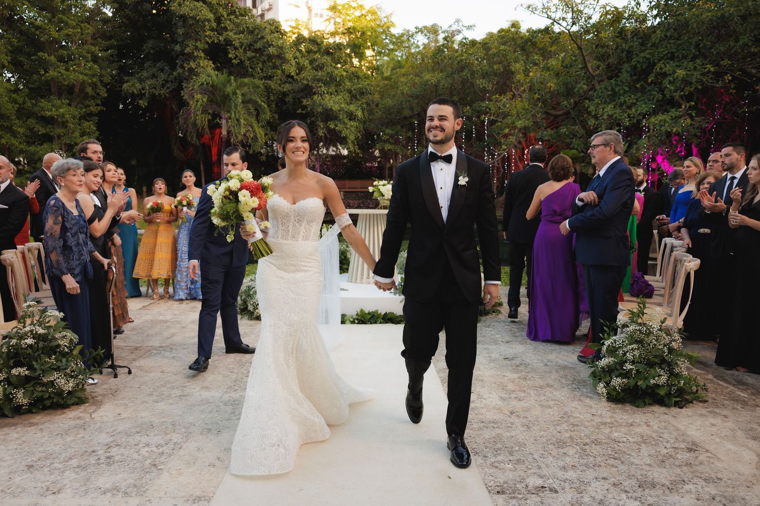 A joyful wedding recessional along a decorated outdoor path with celebrating guests on both sides.