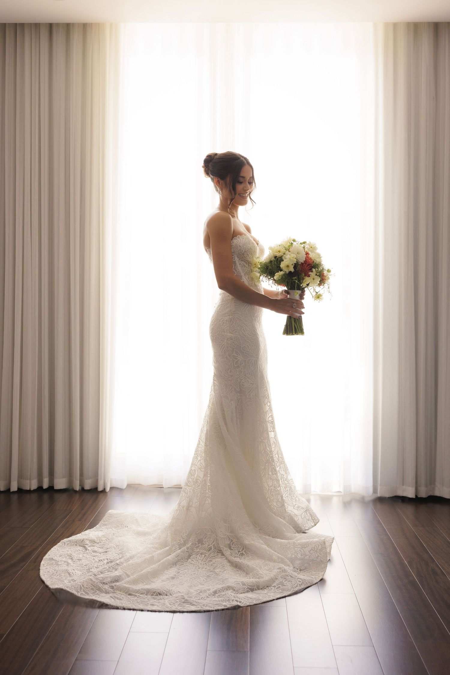 Silhouetted bride in fitted lace gown holds bouquet while standing between white curtains.