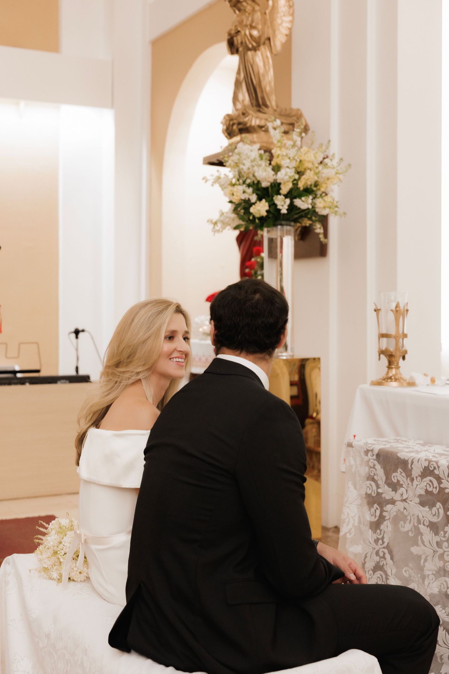 A bride and groom sit together during their wedding ceremony in an elegant church with white floral decorations.