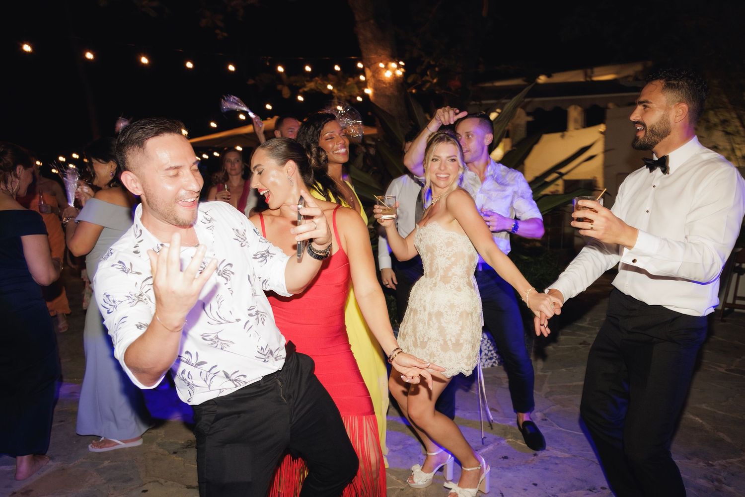 A group of people dancing and having fun at a nighttime wedding reception or party with string lights overhead.