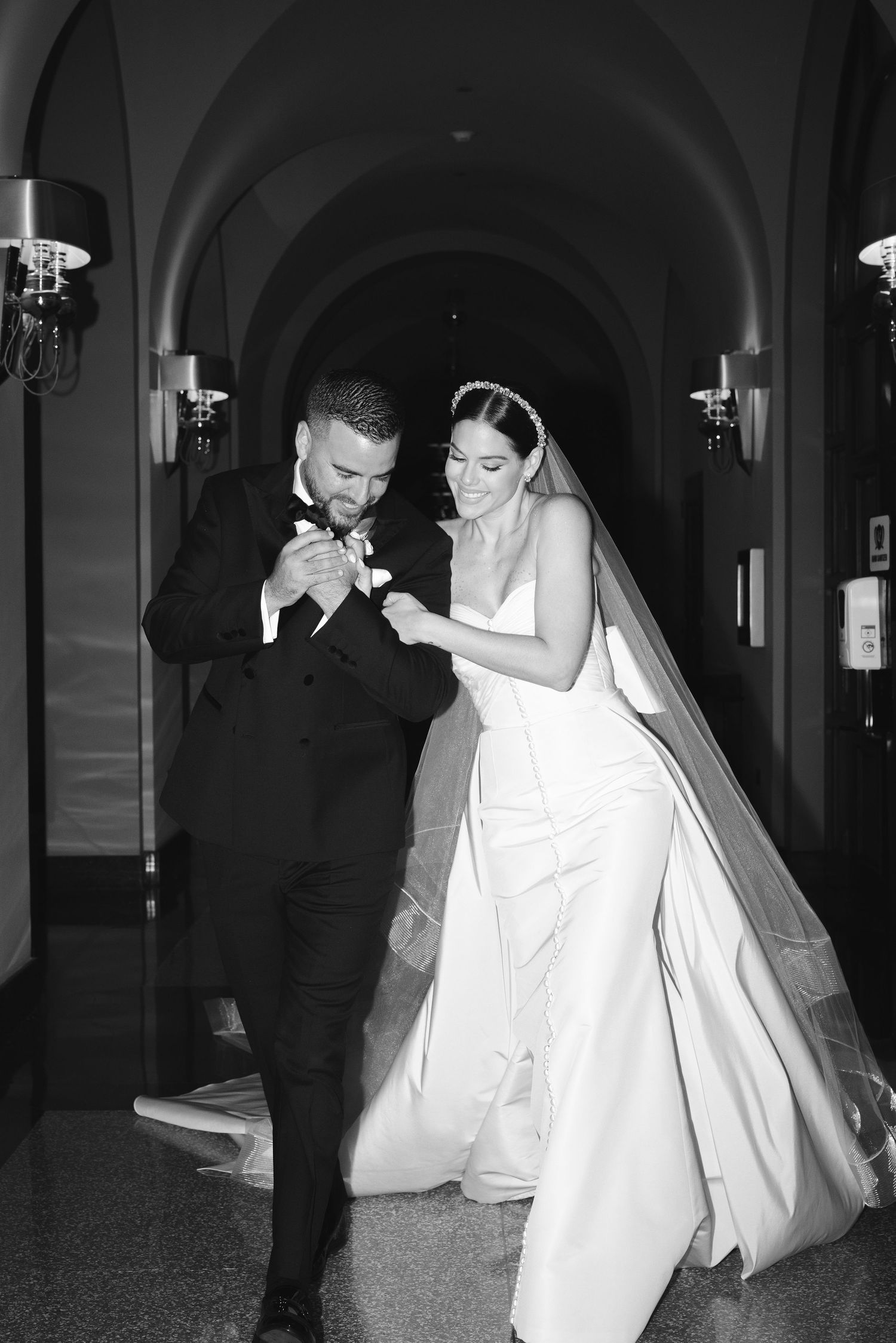 A bride and groom share a joyful moment dancing together in an elegant hallway with arched ceilings and wall sconces.