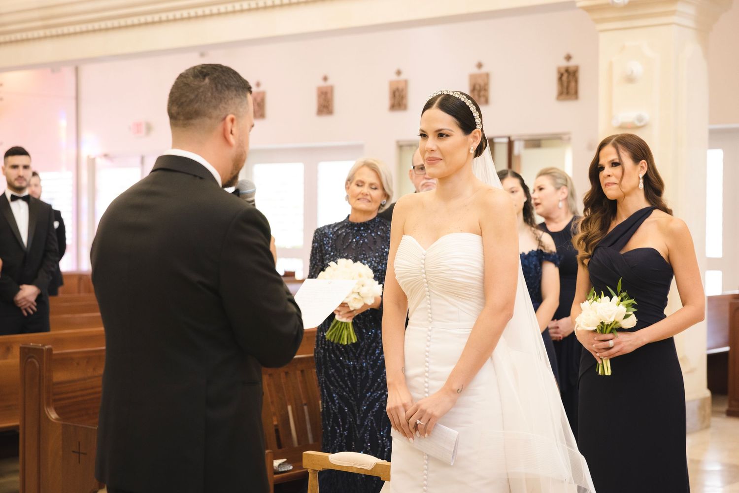 A wedding ceremony takes place inside a bright white church with the wedding party gathered around during the vow exchange.