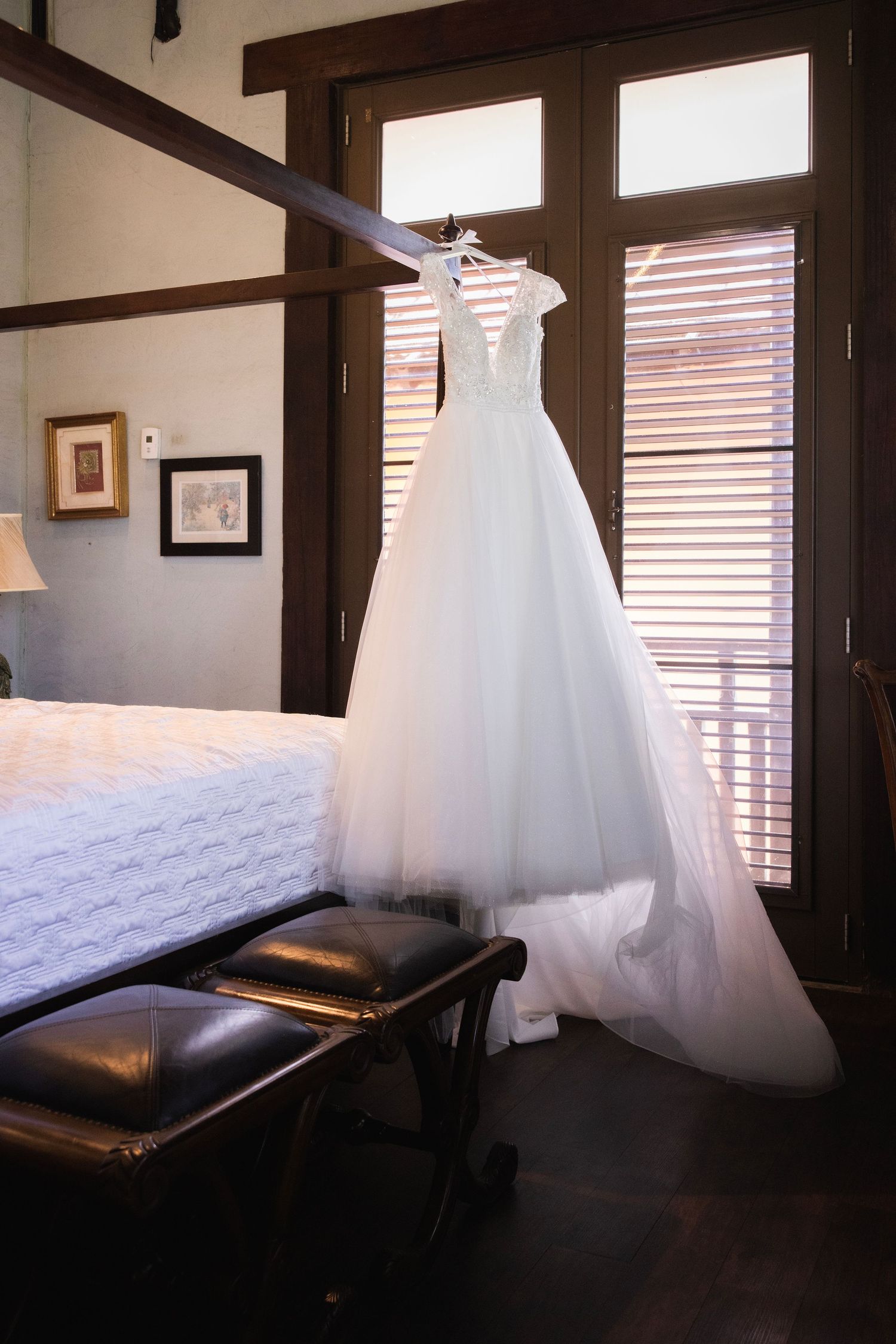 White wedding dress hanging in a sunlit room with dark leather furniture and wooden beams.