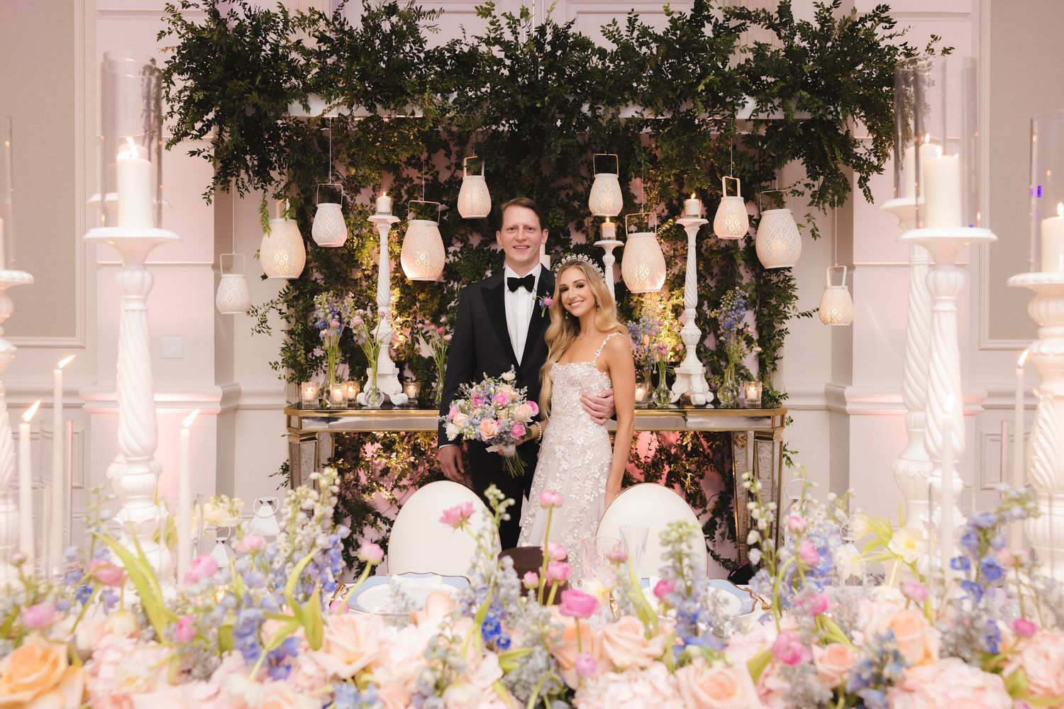A romantic wedding reception setup with hanging lanterns, lush greenery archway and colorful floral arrangements on display.