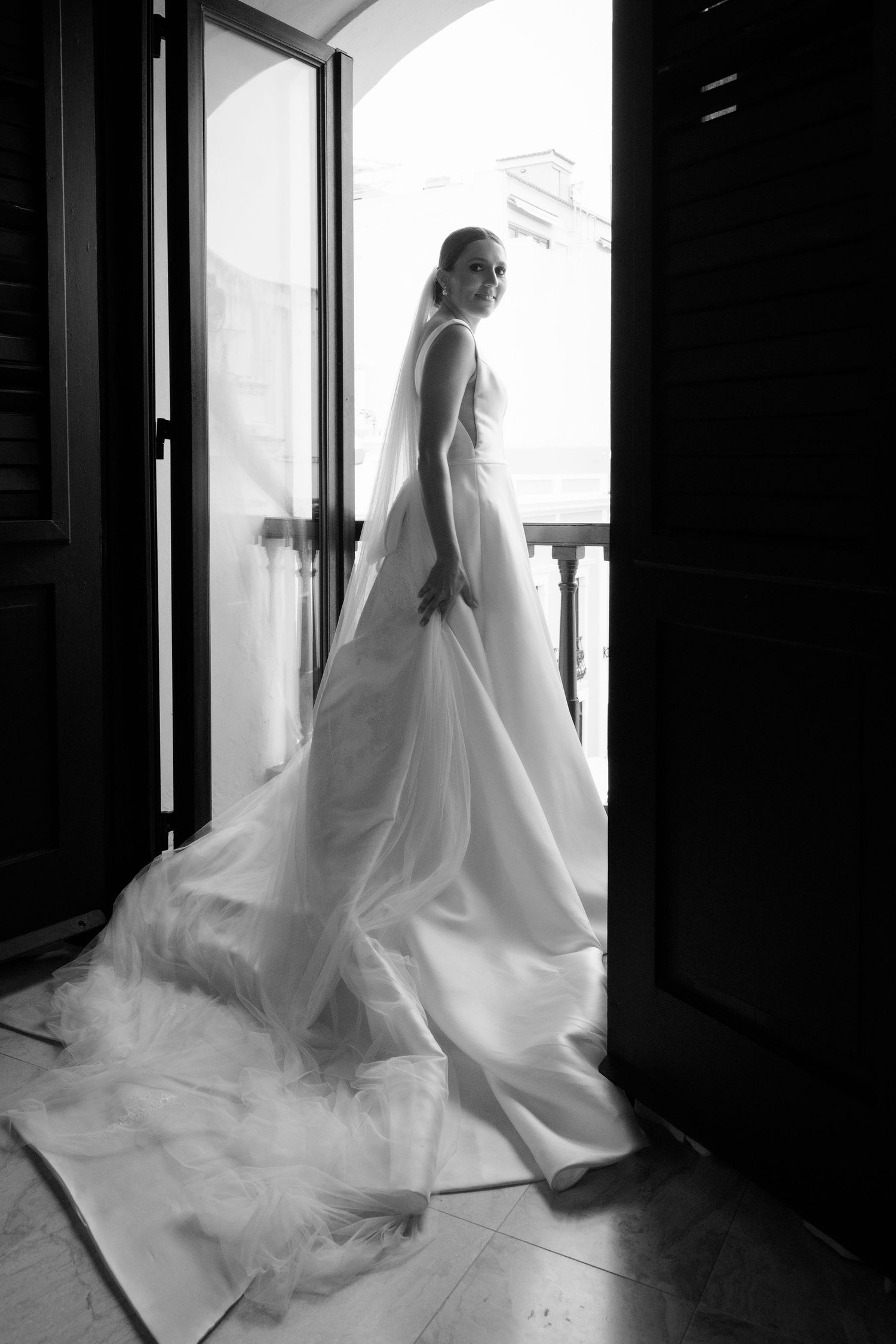 Silhouette of flowing white wedding dress and veil near window in dramatic black and white lighting.