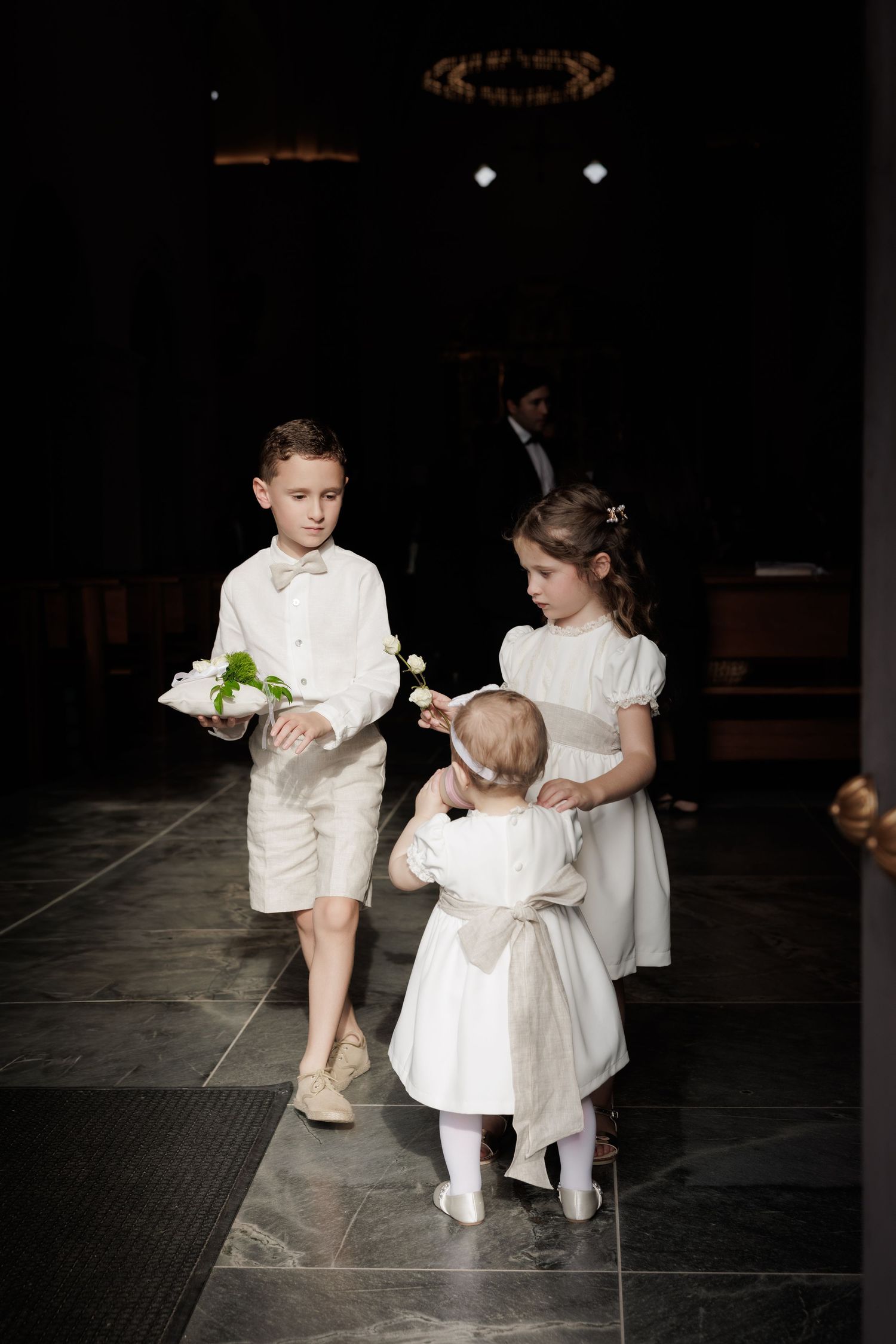 Young children in white formal attire playing and interacting on a dark floor during a special event.