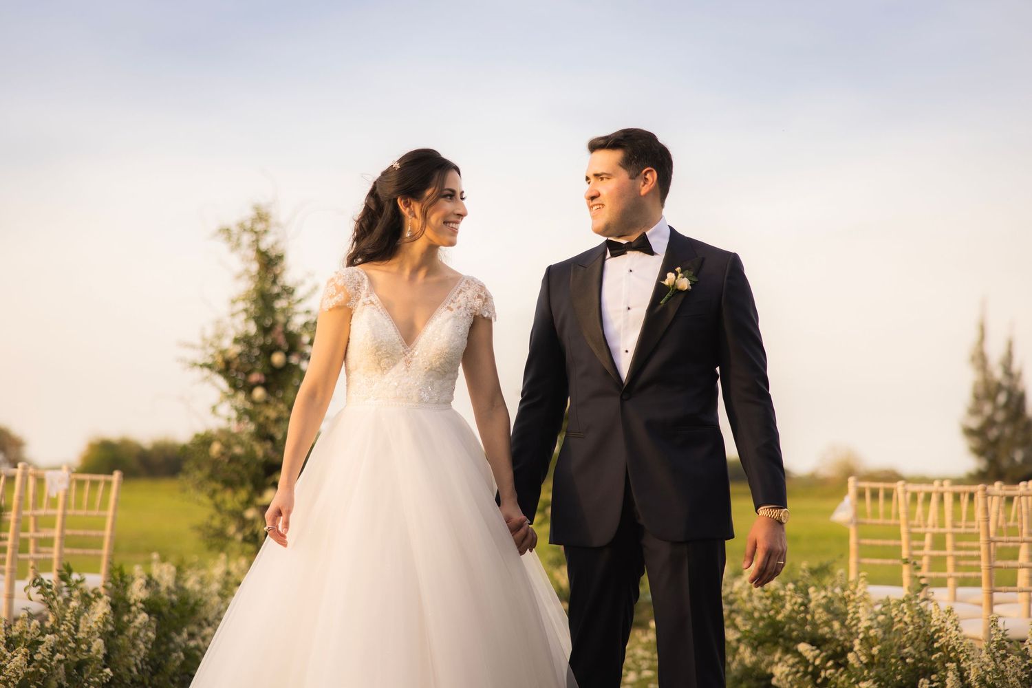 A bride and groom walk hand in hand through a sunlit outdoor vineyard setting at golden hour.