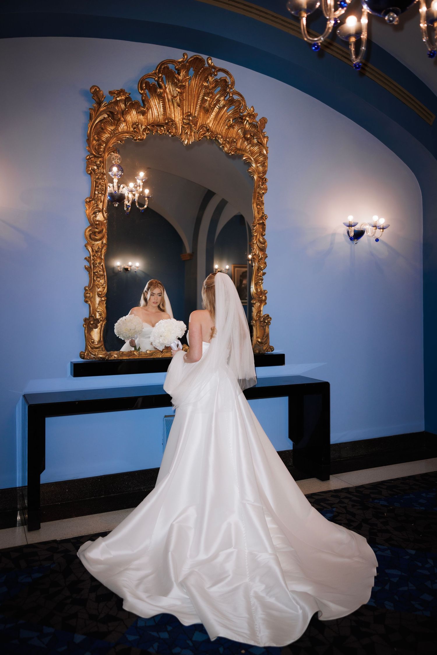 Dramatic bridal portrait in ornate room with gold mirrors and blue walls showing flowing white wedding gown.