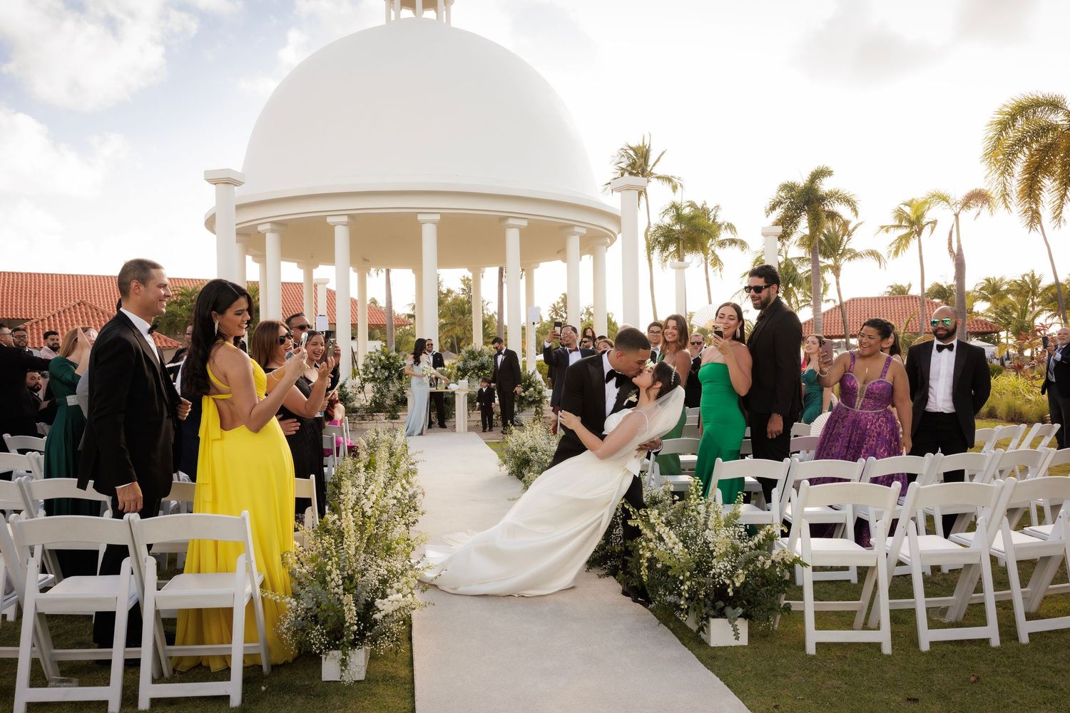 A grand outdoor wedding ceremony with white dome gazebo, palm trees and white folding chairs.