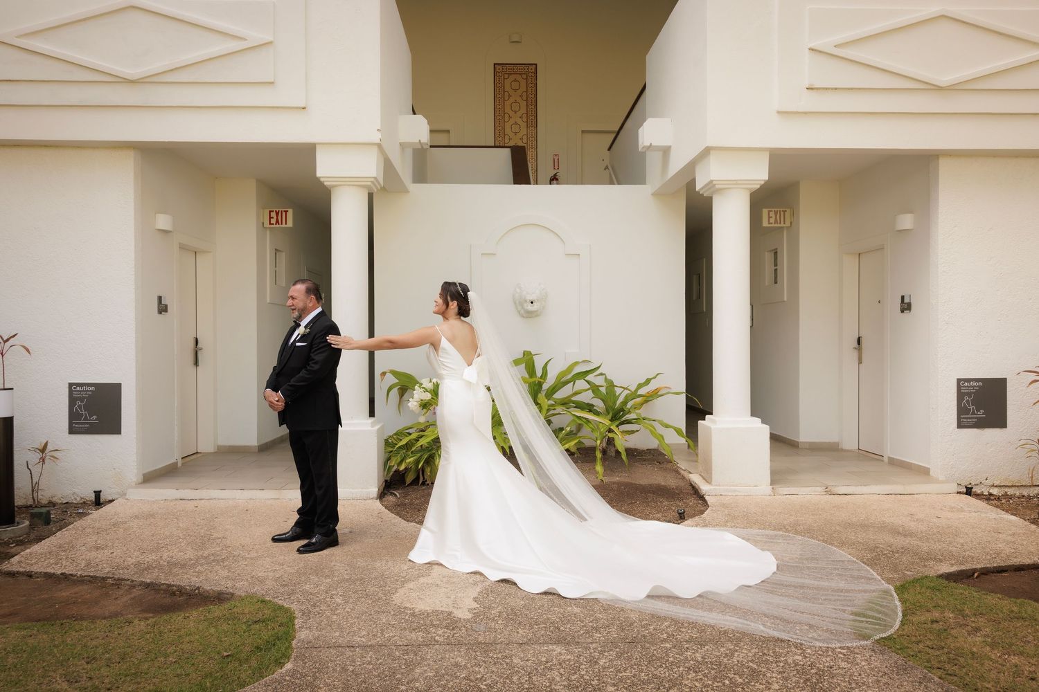 A bride in a flowing white gown with long train touches hands with groom at first look moment between white columns.