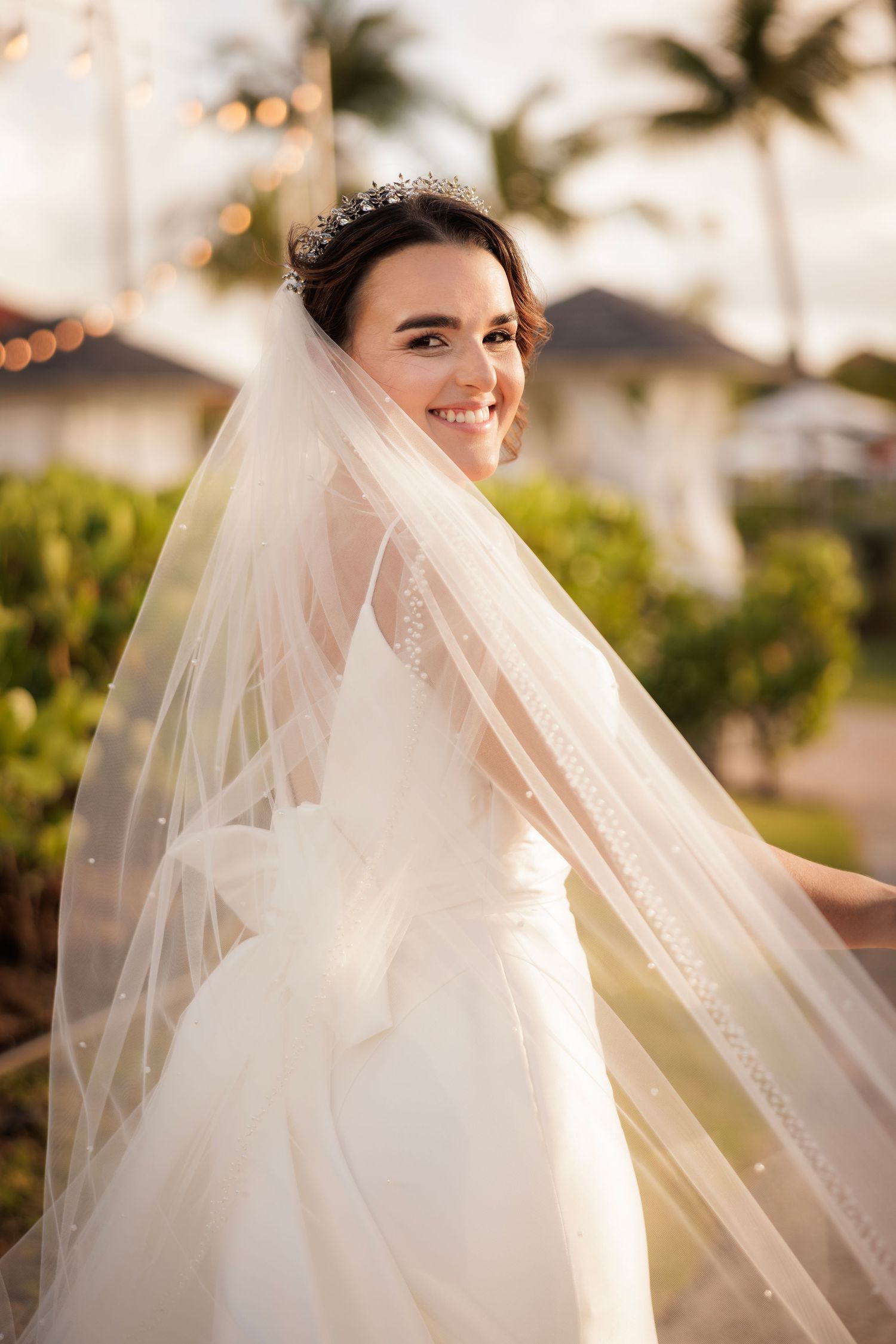 A bride in white wedding dress and veil smiles outdoors with palm trees and sunset lighting in the background.
