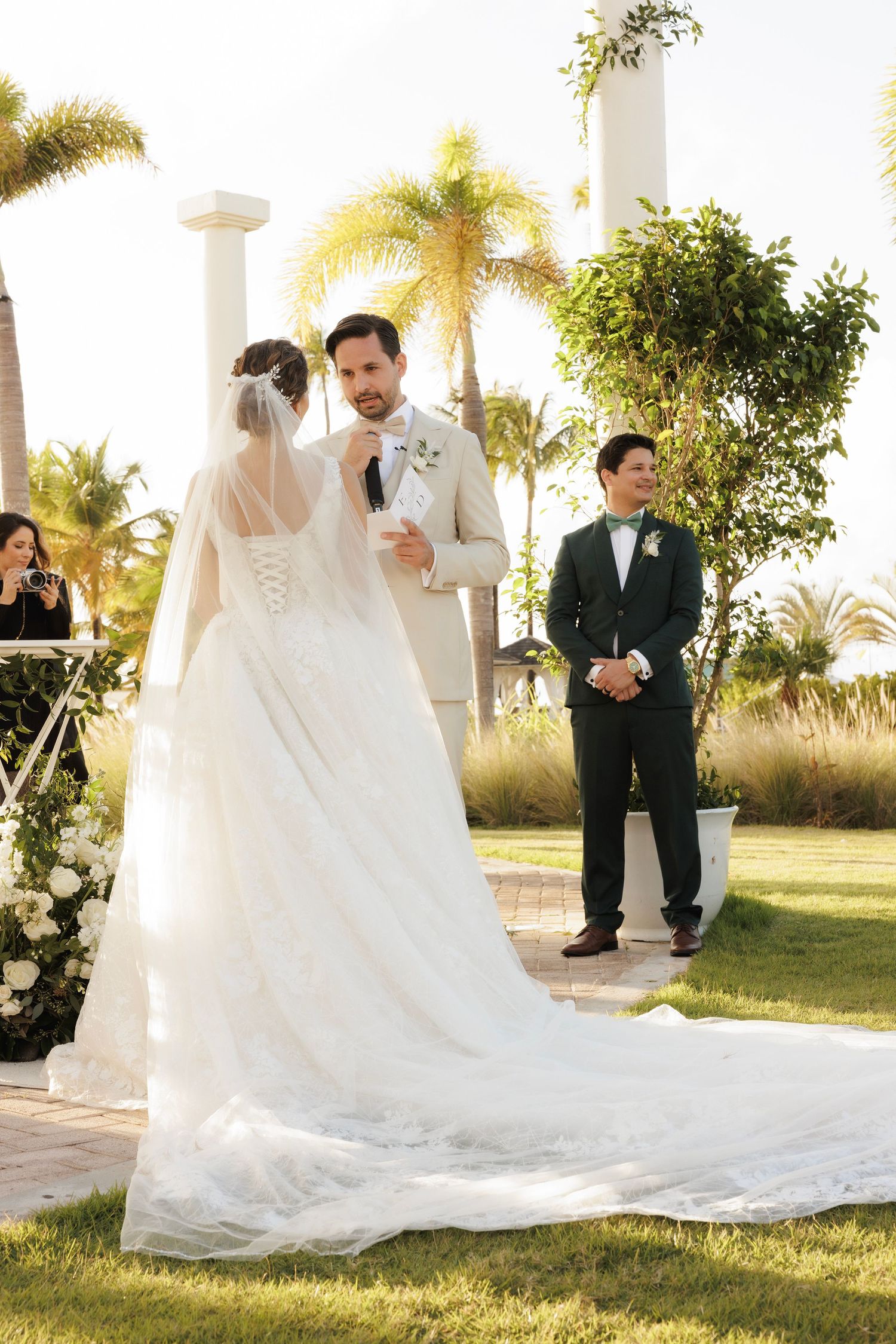 Beautiful outdoor wedding ceremony taking place in a garden setting with palm trees and white pillars at sunset.