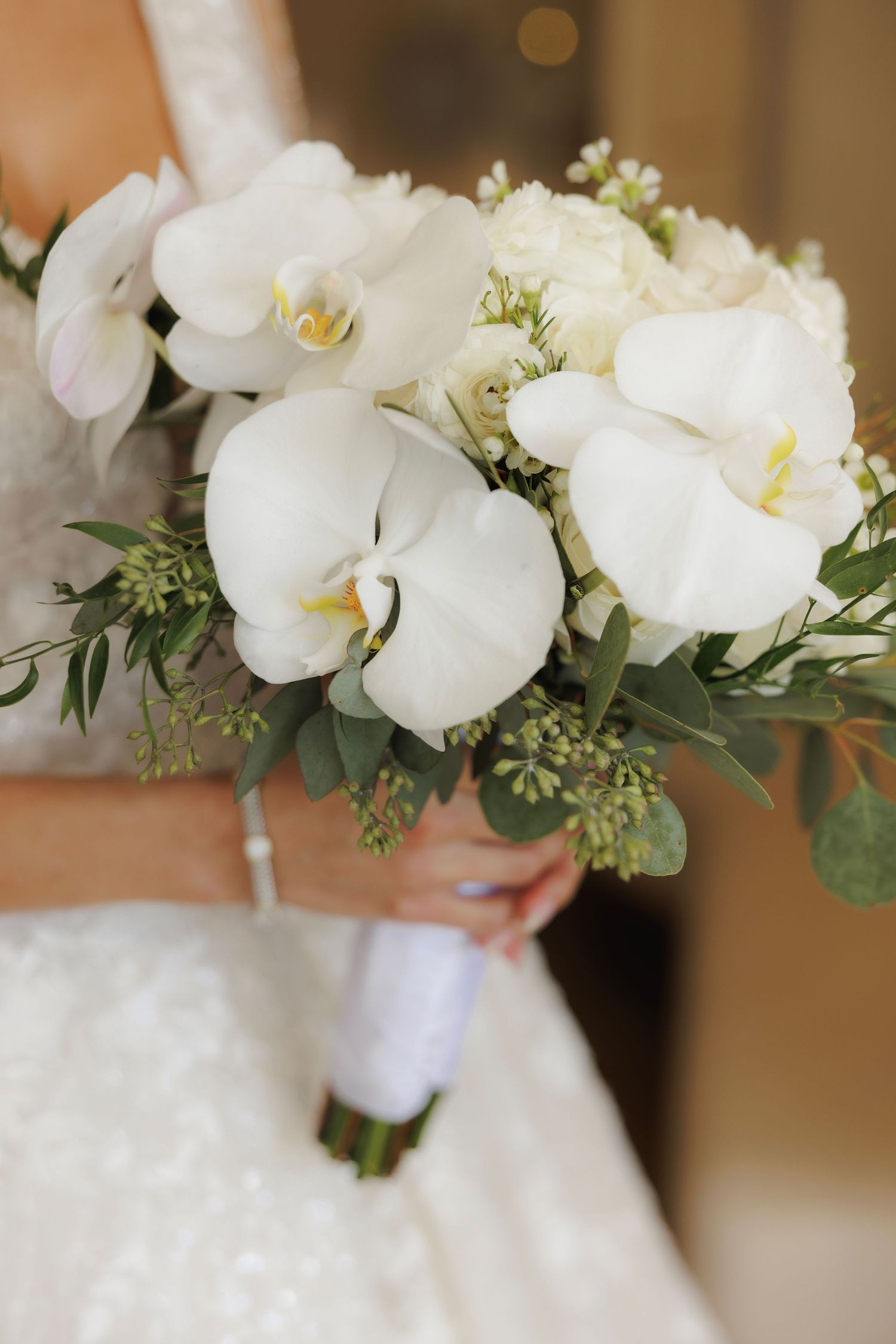 Close-up view of elegant white orchid wedding bouquet with delicate greenery accents and soft blurred background.