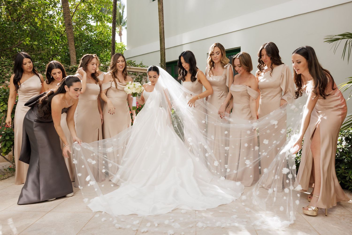 A group of bridesmaids in blush pink dresses gather around a bride in a white gown during an outdoor wedding celebration.