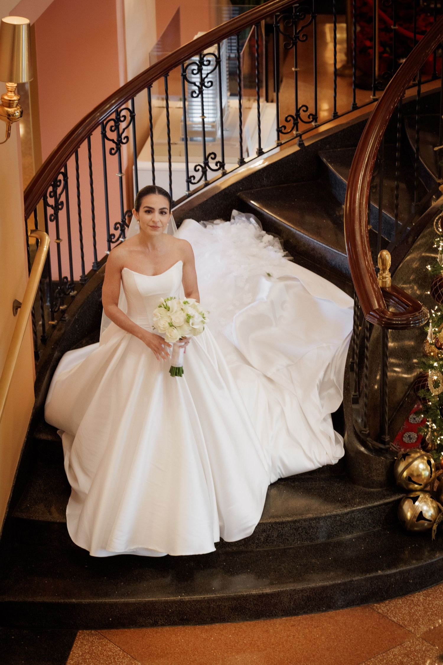 A bride in a voluminous white wedding gown poses elegantly on a curved staircase with ornate iron railings.