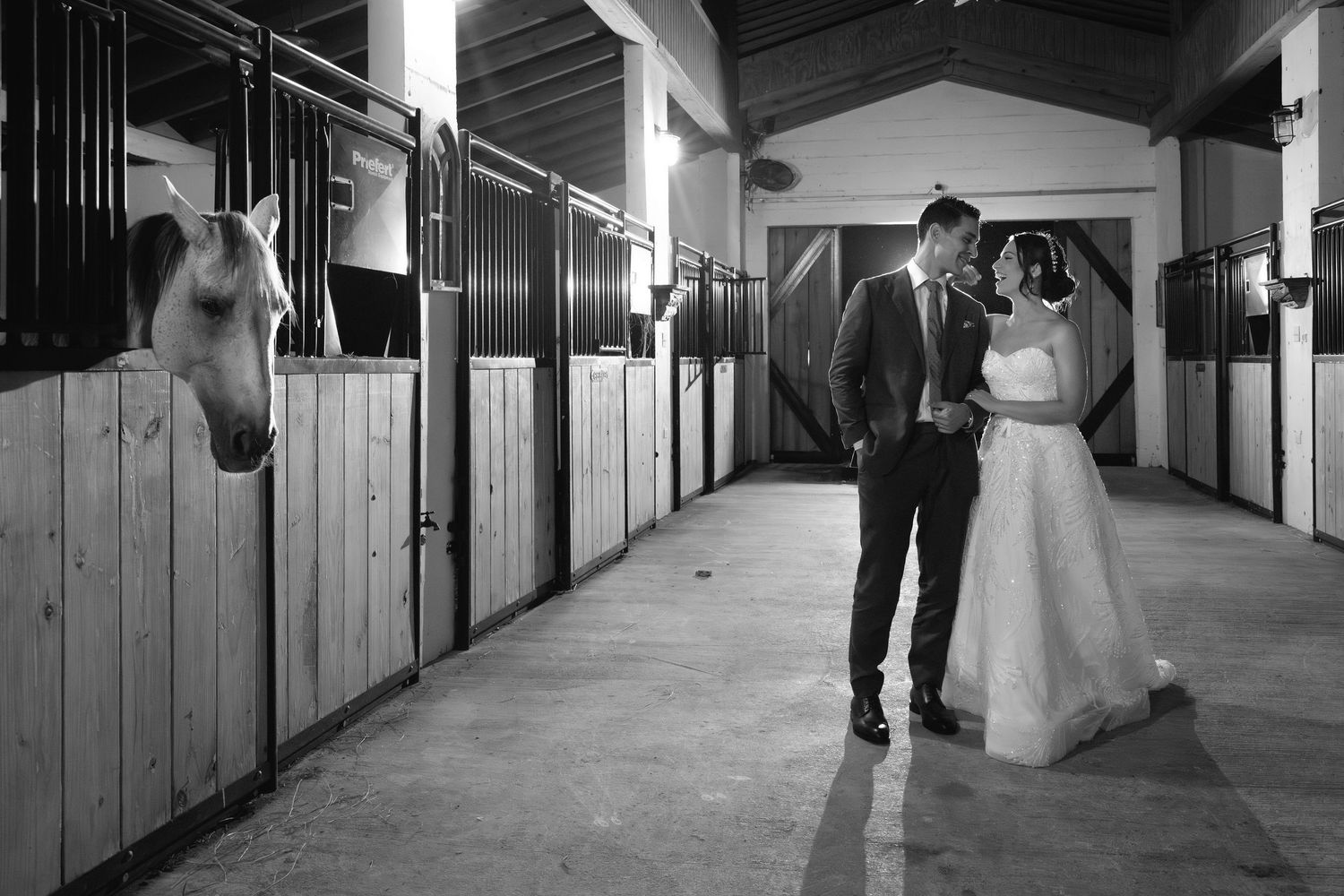 A couple walks through a rustic stable corridor illuminated by overhead lights casting long shadows.