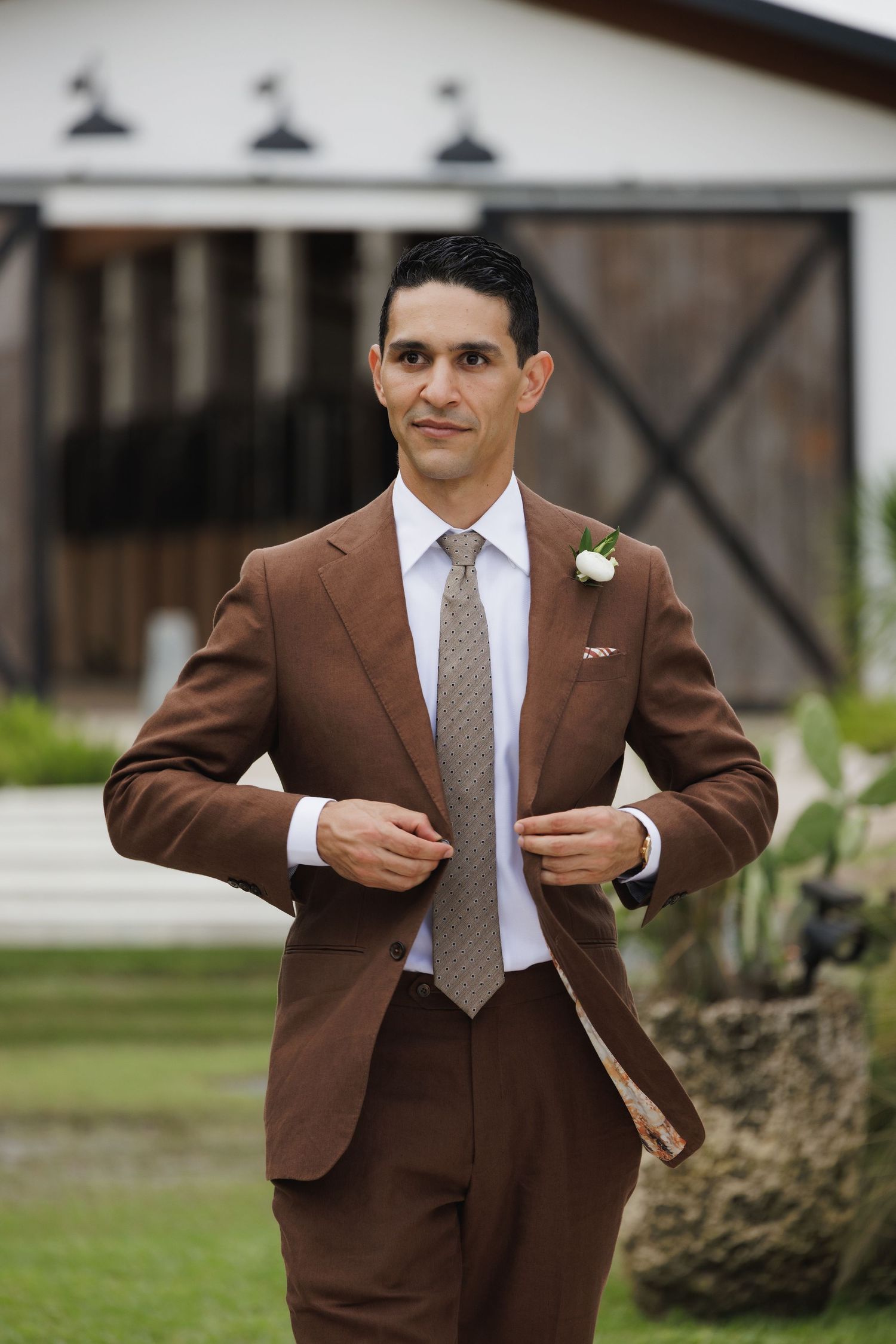 A well-dressed figure in a tailored brown suit adjusts their jacket outdoors.