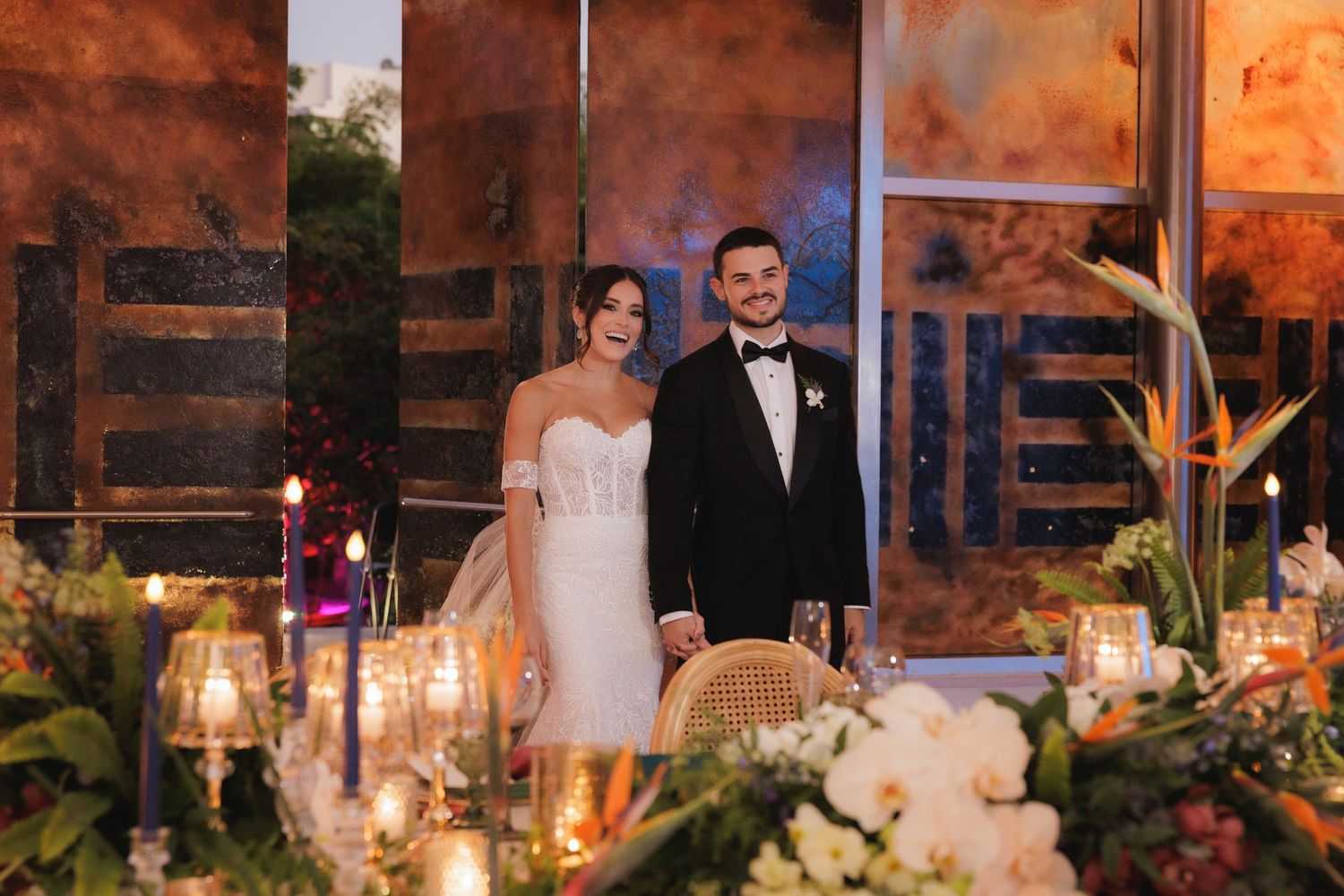 A couple stands behind an elegant reception table decorated with glowing candles and lush floral arrangements.