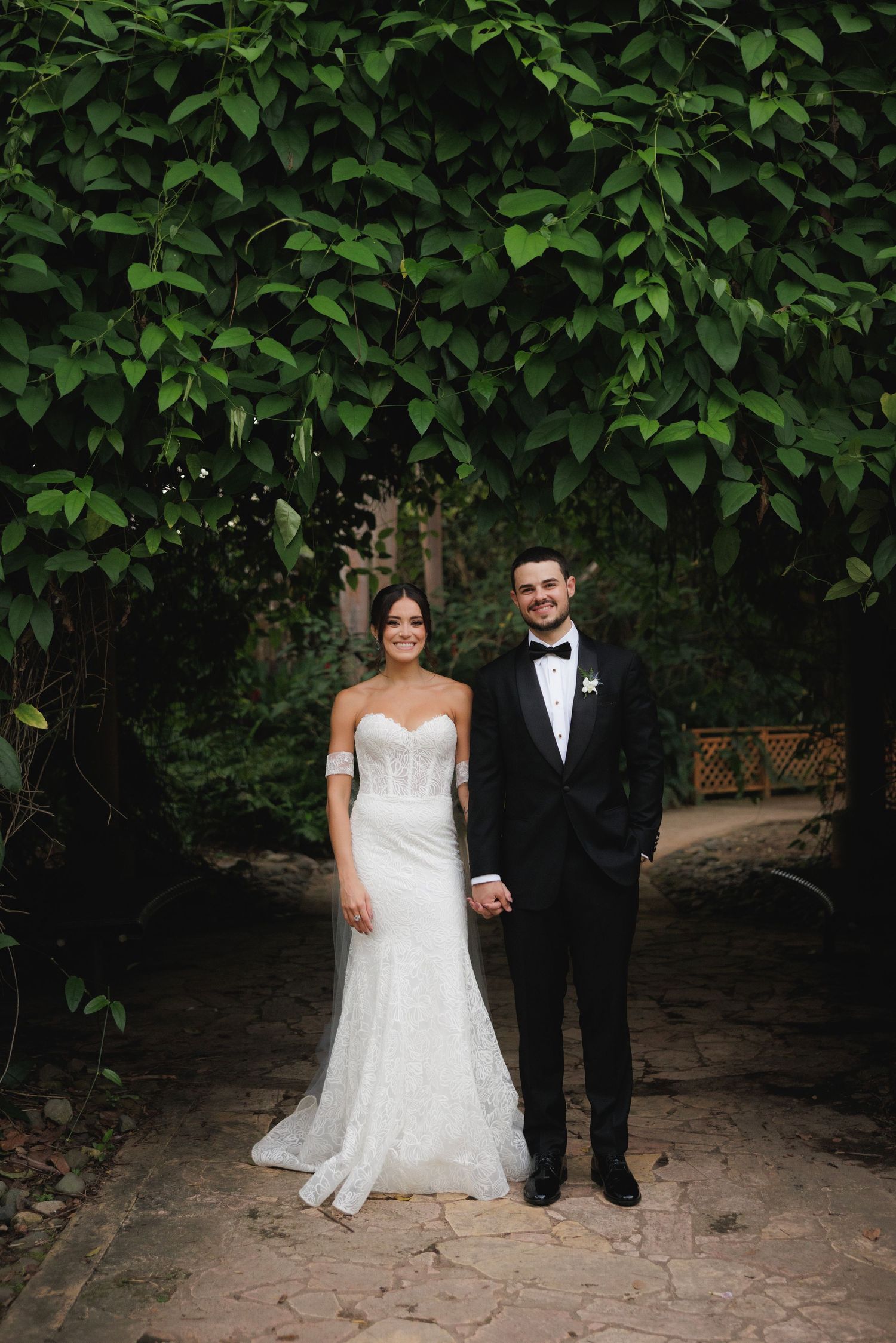 Wedding couple poses against backdrop of lush green foliage in garden setting.