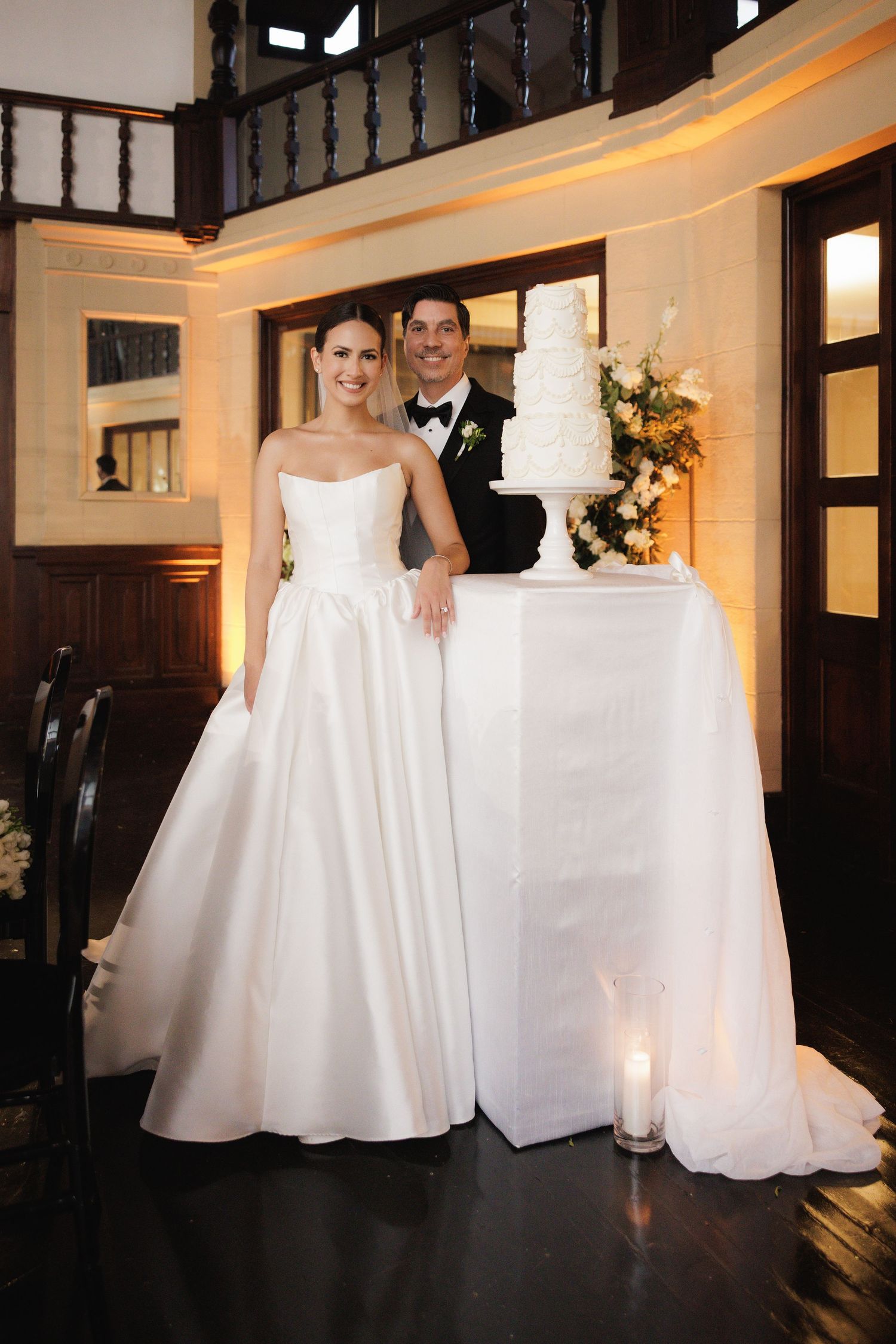 Wedding couple poses in an elegant indoor venue with dark wood railings and white gowns creating contrast.