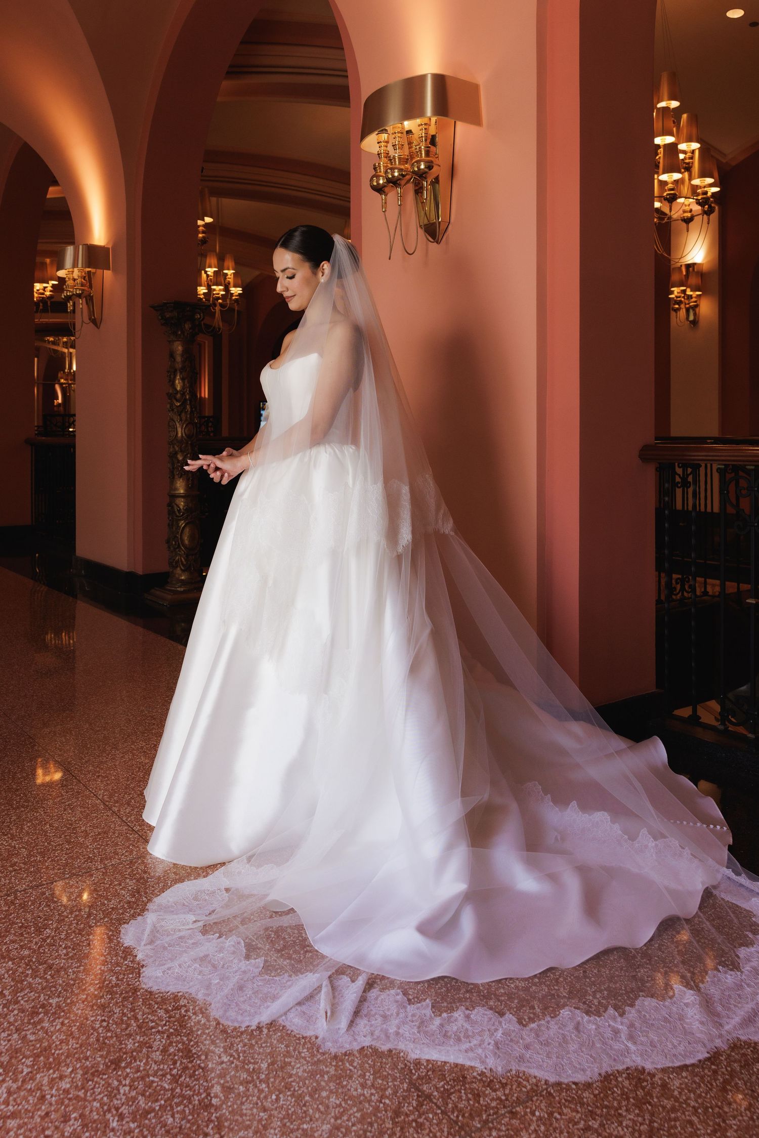A flowing white wedding gown with a dramatic train photographed in an elegant ballroom with warm lighting and chandeliers.
